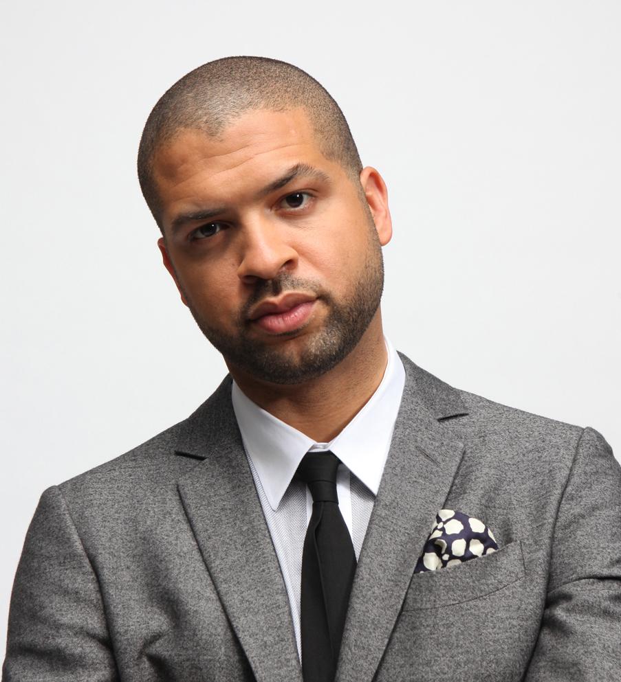 [Image description: Photo of Jason Moran standing against a white background. He is wearing a gray suit, white collared shirt, and black tie.]