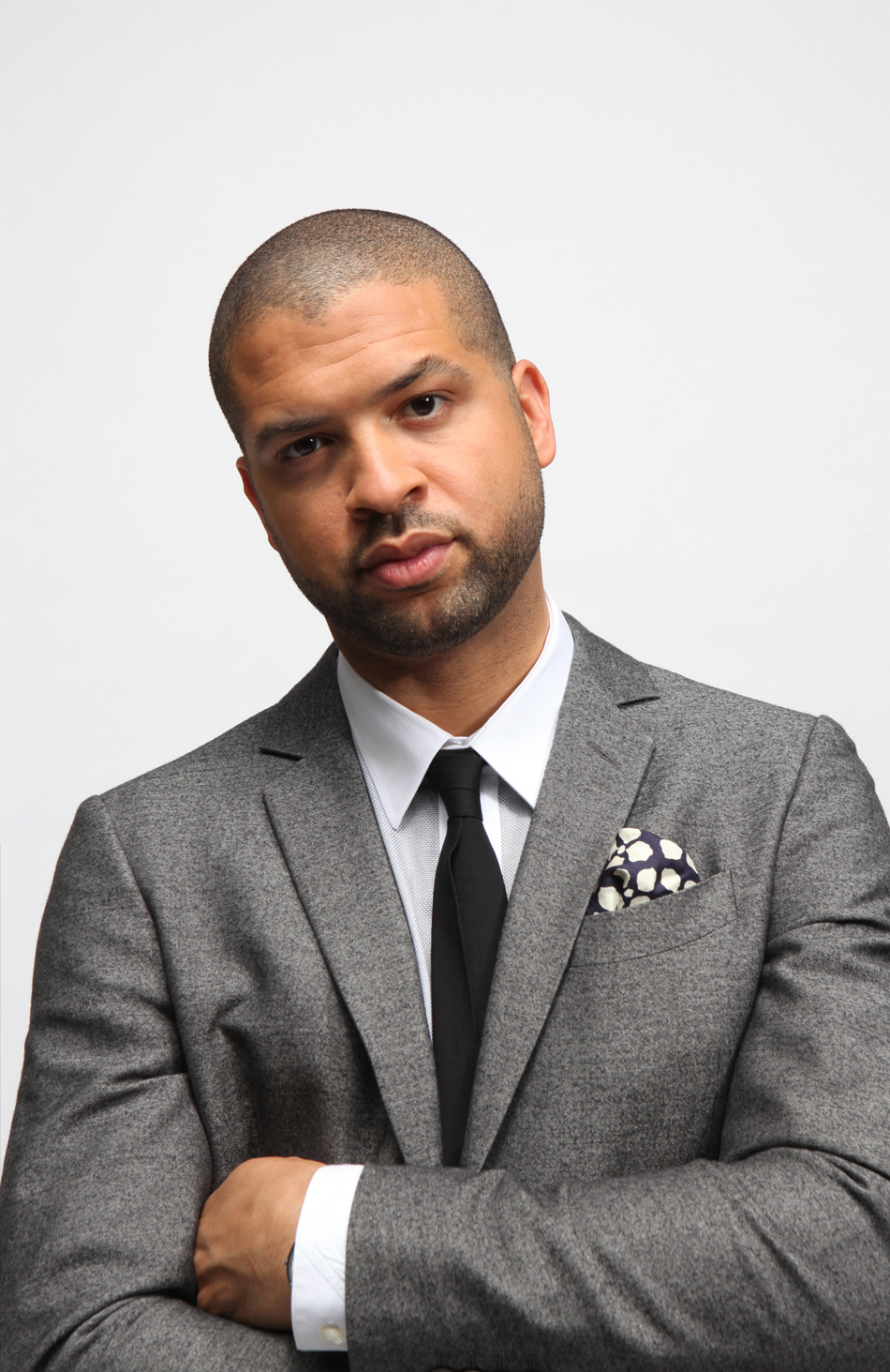 [Image description: A photo of artist Jason Moran. He stands with his arms crossed looking directly into the camera. He wears a gray suit jacket, white collared shirt, black tie, and black and white pocket square.] 