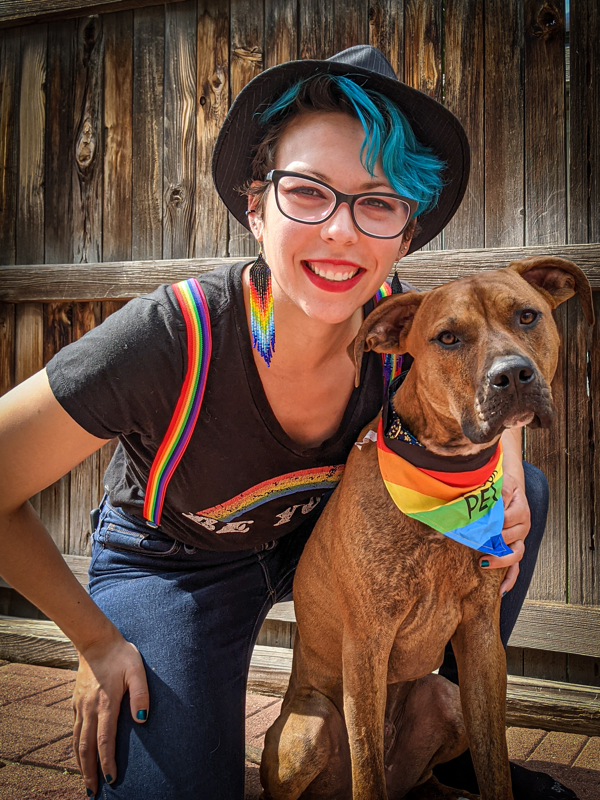 [Image description: Photo of Jacenta L. Irlanda crouched in front of a wooden fence. She is smiling at the camera with a large mutt dog. She is wearing a black t-shirt, blue jeans, rainbow suspenders, and a black fedora atop her cool blue-dyed hair. The dog is wearing a rainbow bandana around its neck.]
