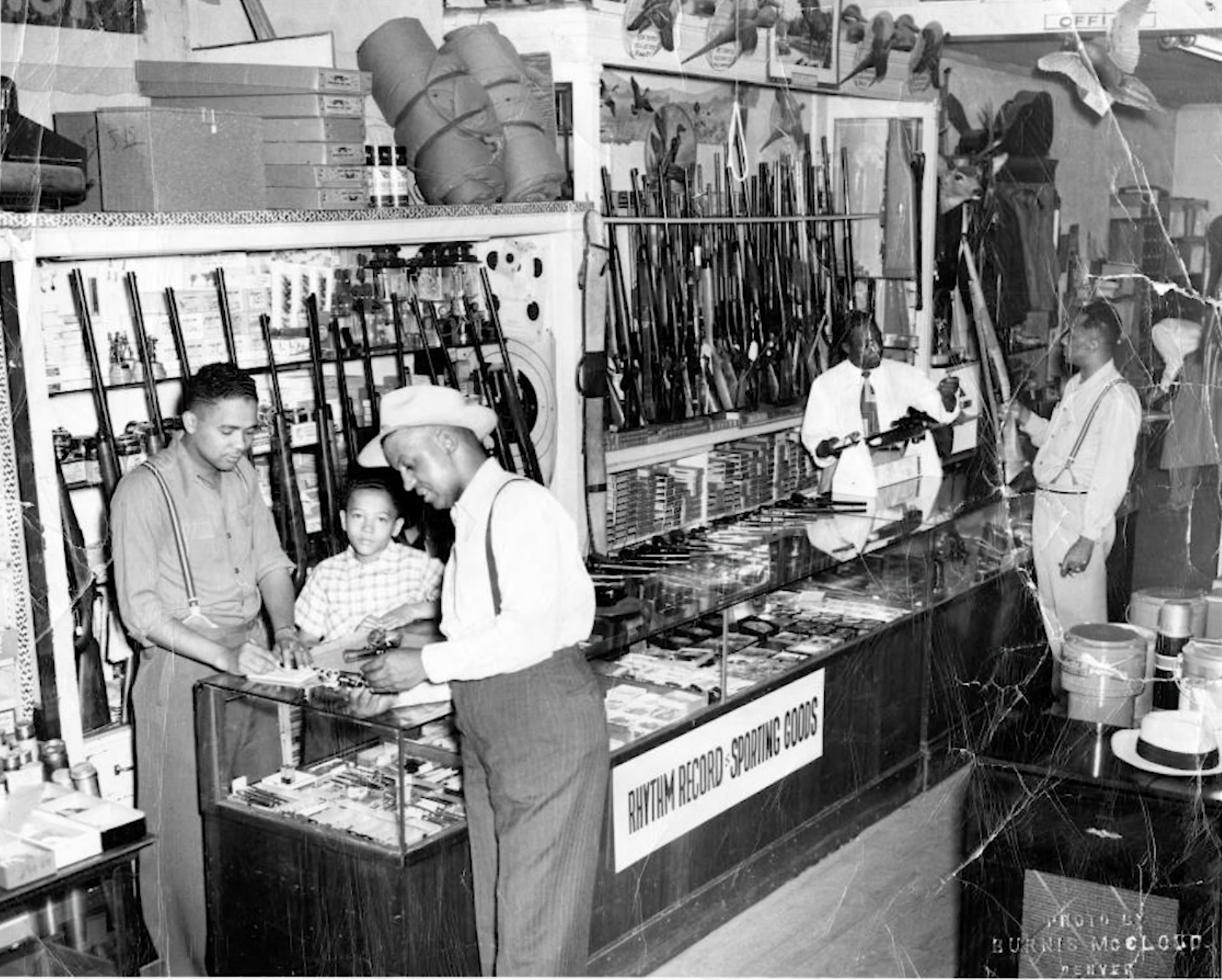 [Image Description: Black and white photograph of a shop in 1940's Denver. Rifles line the walls and Black men are seen standing and discoursing.]