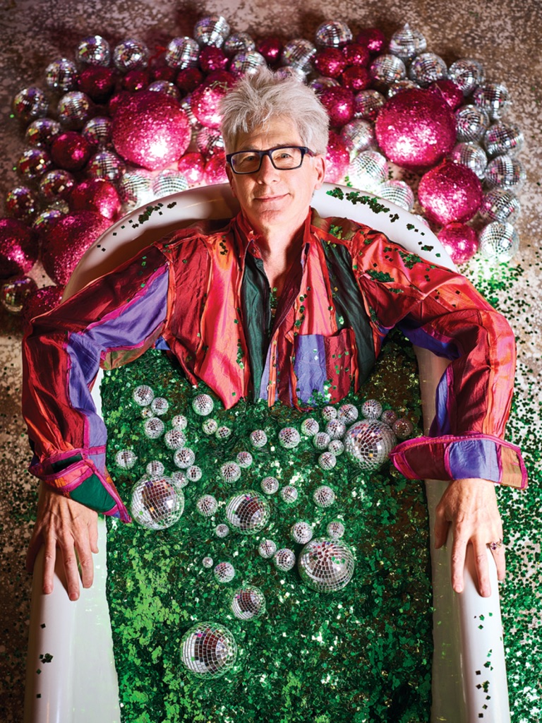 [Image description: A photo of Lonnie Hanzon. The photo is taken with a bird's eye view looking down at Lonnie in a bathtub looking up at the camera. The bathtub is filled with green glitter and silver Christmas tree ornaments and the floor is covered in magenta and silver ornaments. Lonnie has short white hair, glasses, and is wearing a flamboyant red and purple long-sleeved, collared shirt.]