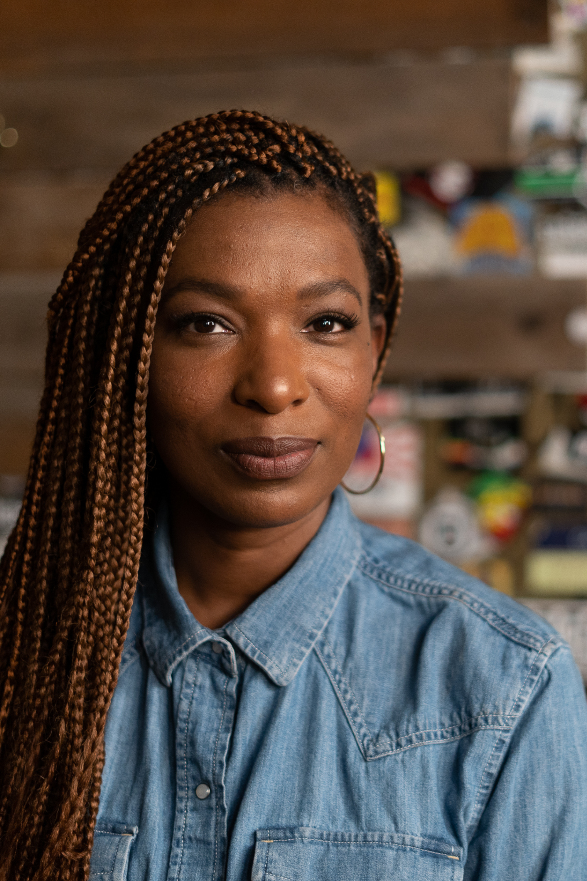 [Image description: Photo of comedian Janae Burris. Janae is sporting a denim shirt and her long brown hair is in braids. She is standing in front of a brick wall with stickers.]