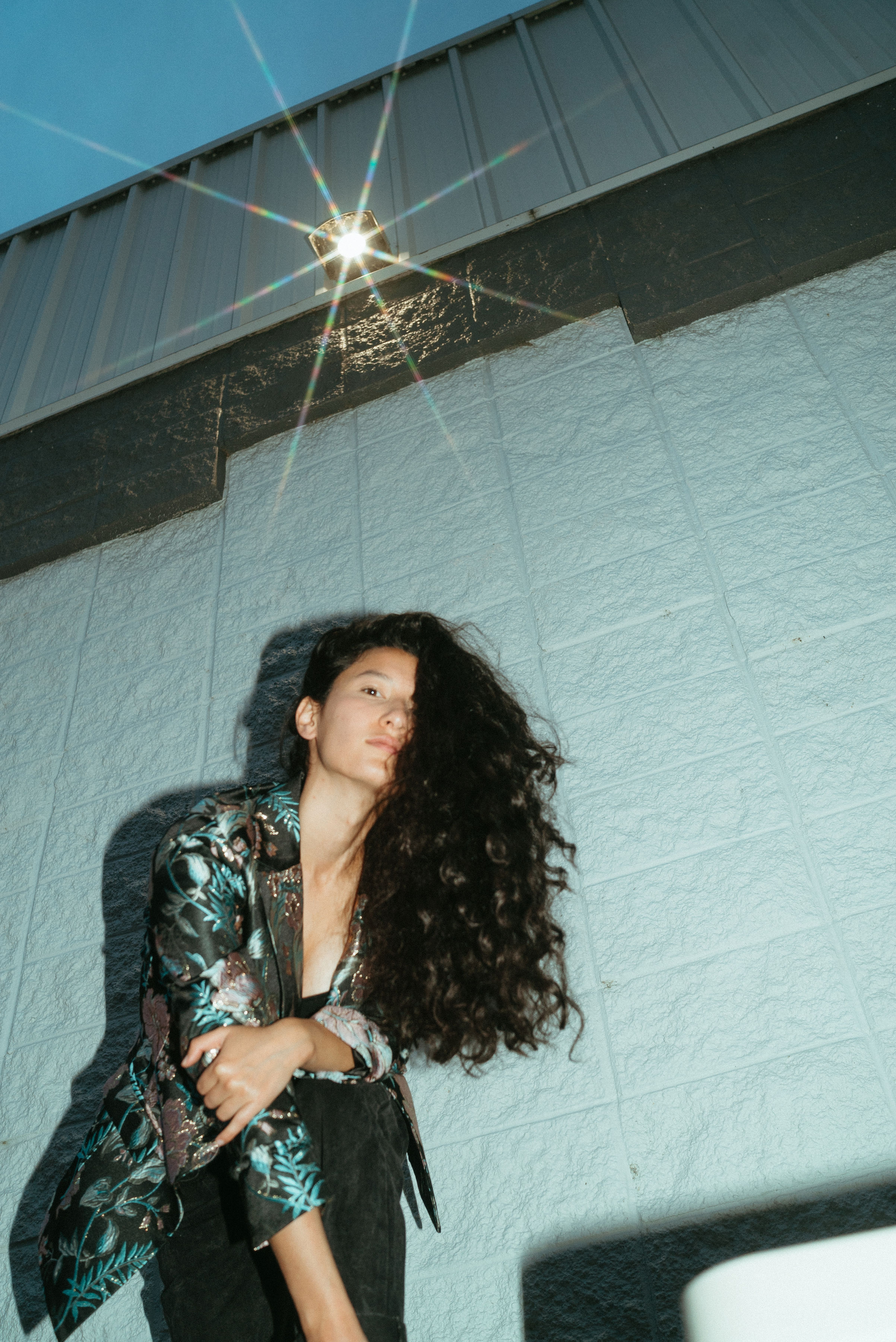 [Image description: Photo of ZEMBU standing in front of an industrial white and gray wall. A small light is above her casting a starburst light reflection. ZEMBU is wearing an amazing black blazer with teal and soft pink floral brocades. She has long, dark, curly hair.] 