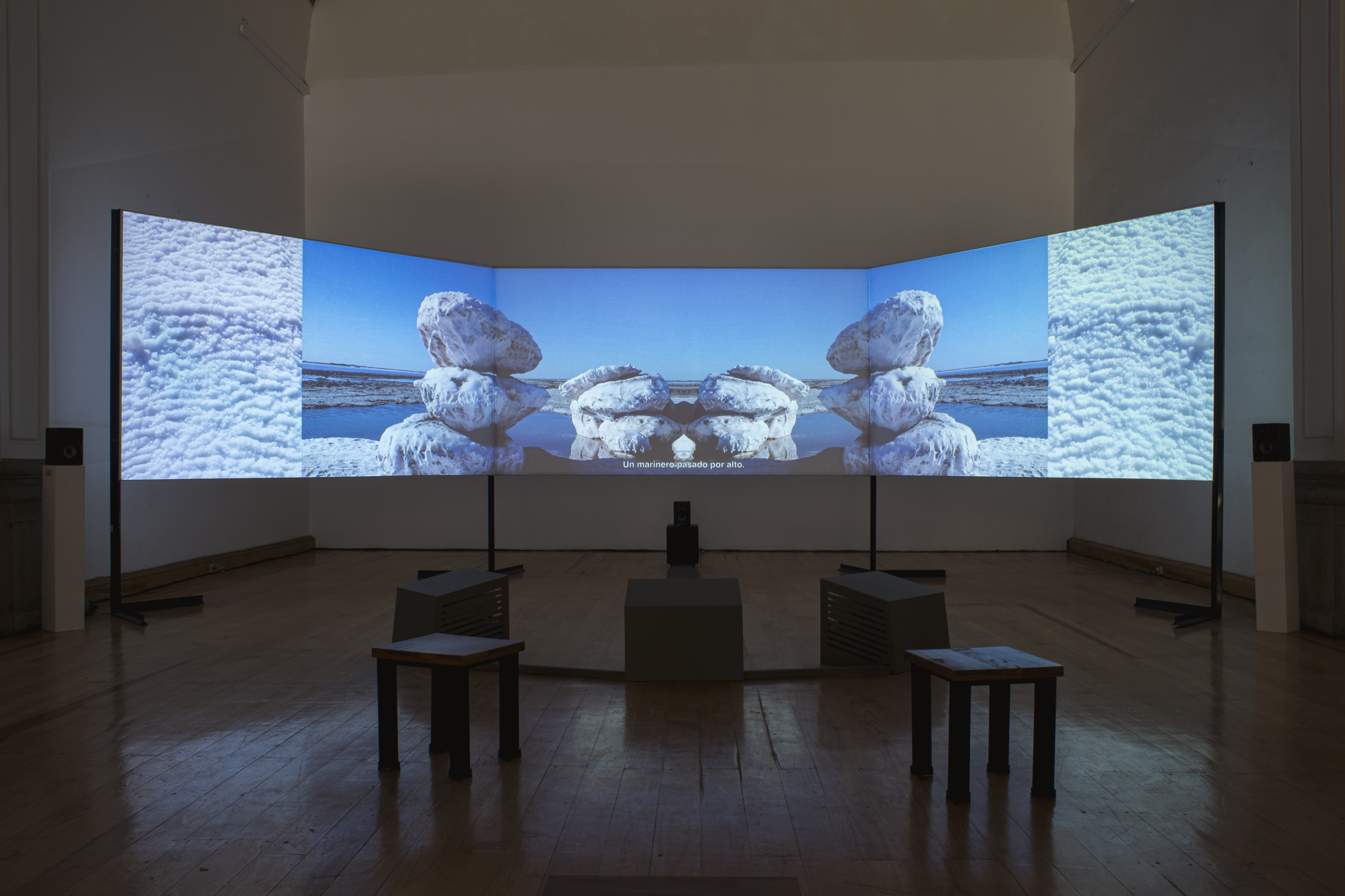 Large three-channel screen in a dark room. The images on the screen feature a body of water, snow, and what looks like large rocks stacked on top of one another. 