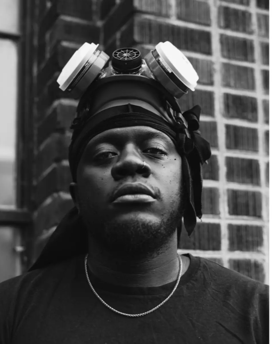 A black and white portrait of Blake Jackson posing in front of a brick wall with a stoic expression on my face and a respirator mask on top of his head.