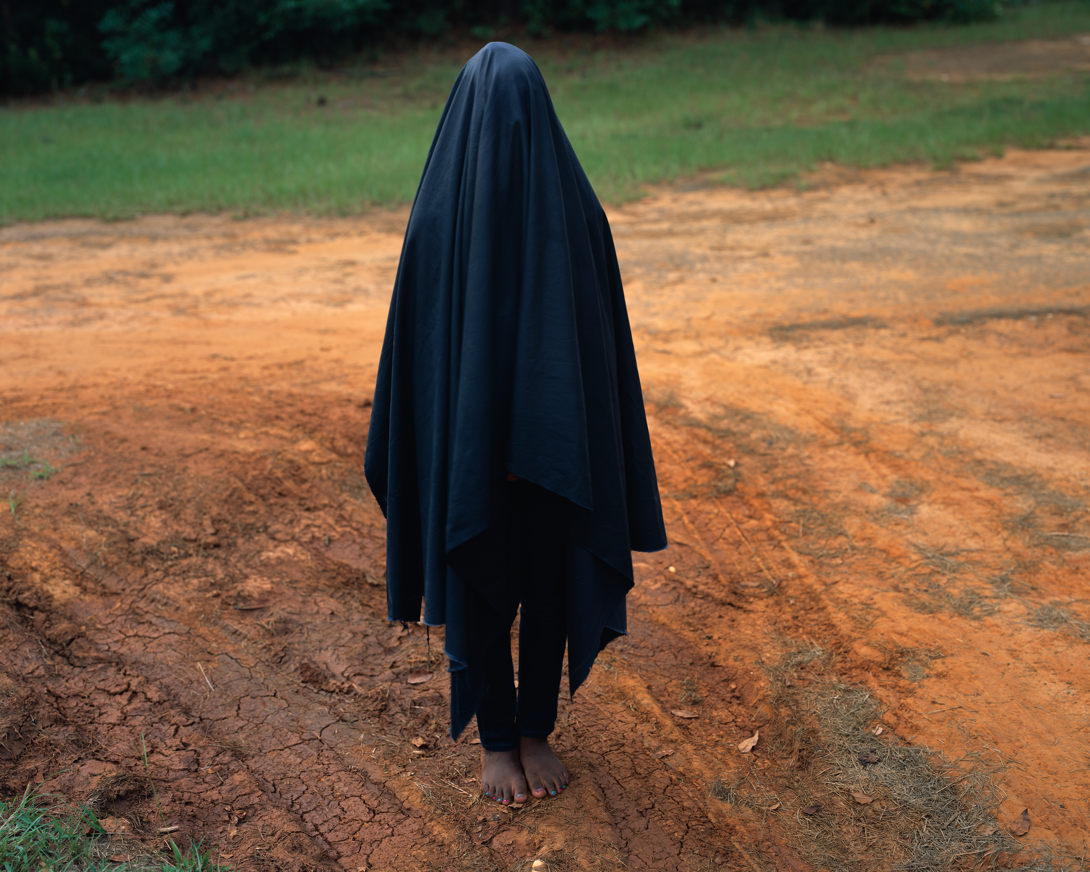 Figure standing barefoot in red dirt, covered in a long black sheet-like material.