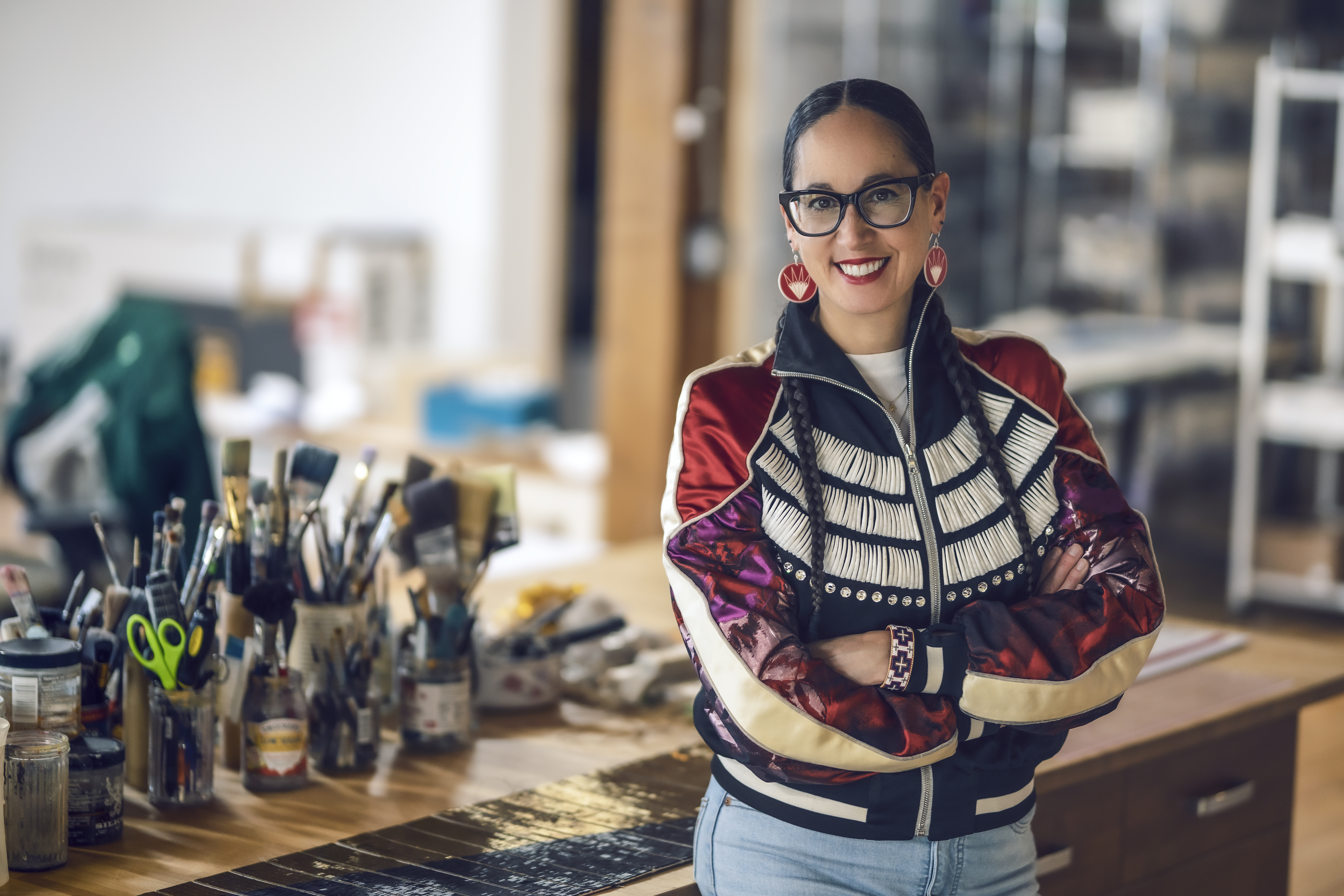  Portrait of artist Dyani White Hawk in her studio, standing in front of a desk with various containers on it that are filled with brushes and other tools. Dyani is sporting a colorful patterned bomber jacket, jeans, big red earrings, two long braids, and glasses with black frames. 