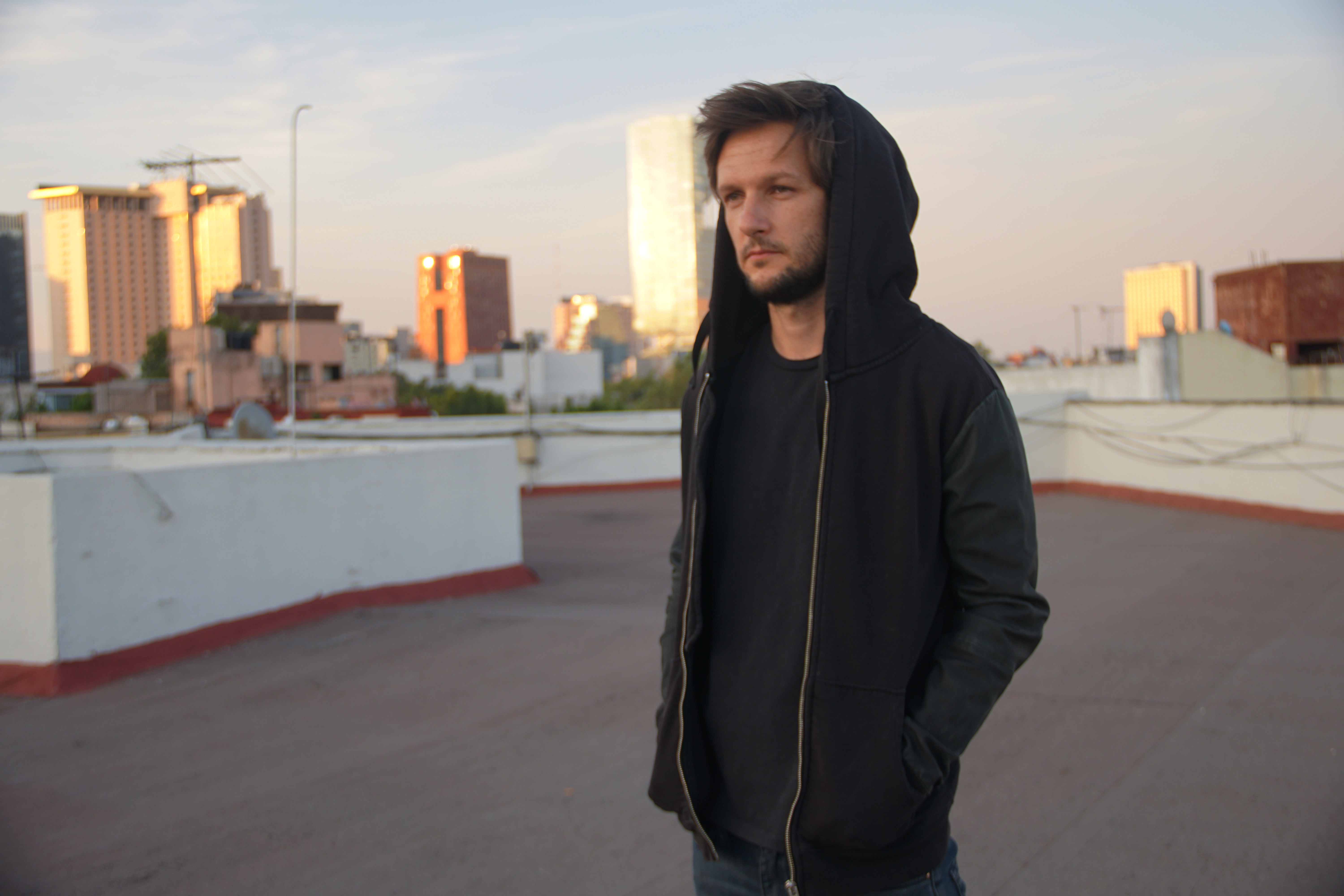 Portrait of Diego Gerard Morrison on a rooftop wearing a black hoodie with the hood pulled up over his head looking off into the distance