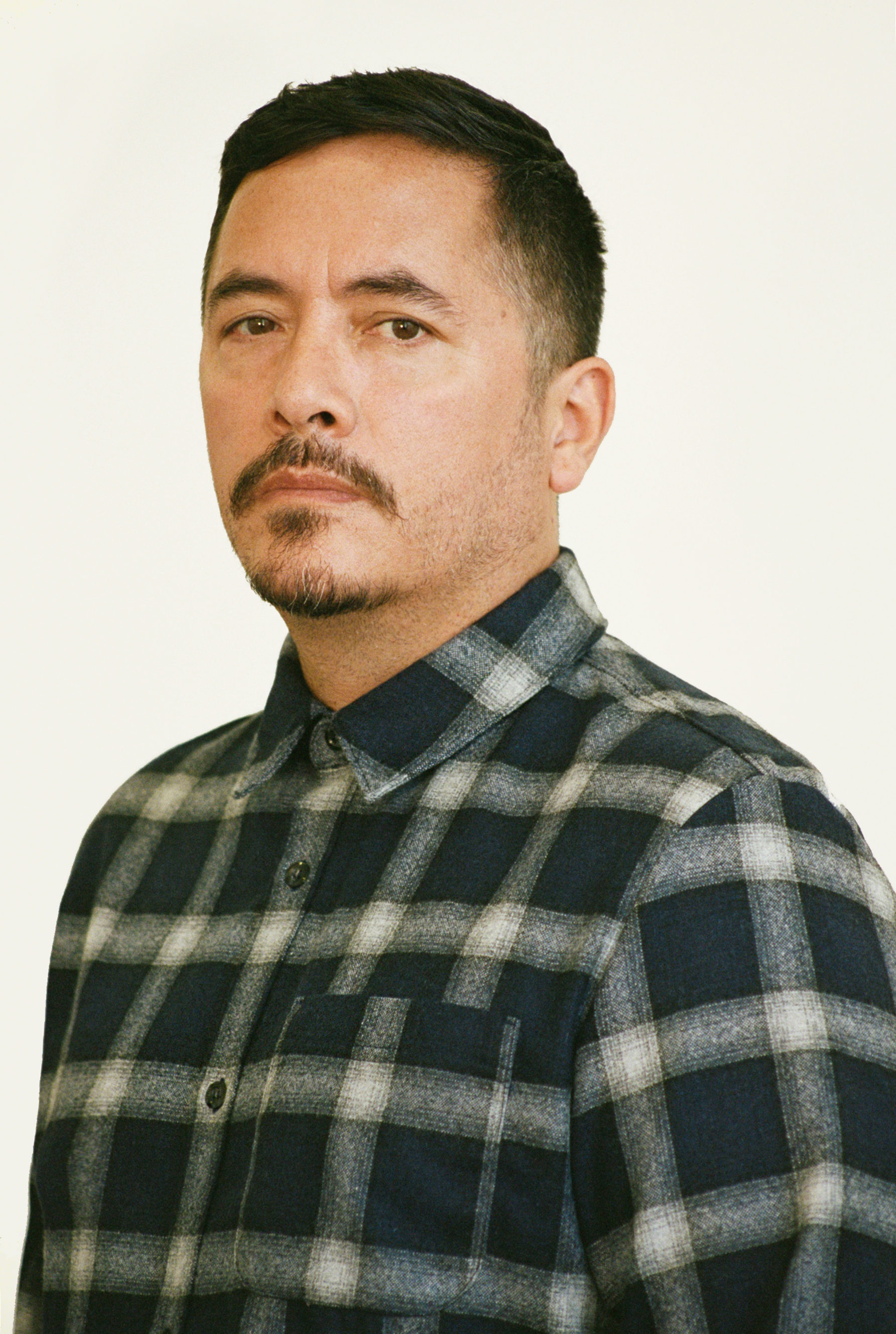 Portrait of artist Eamon Ore-Giron standing in front of an off white, almost cream colored wall. Eamon is sporting a blue and white button-up flannel and has a serious look on his face. His black hair is cut short and he is sporting a mustache and goatee. 