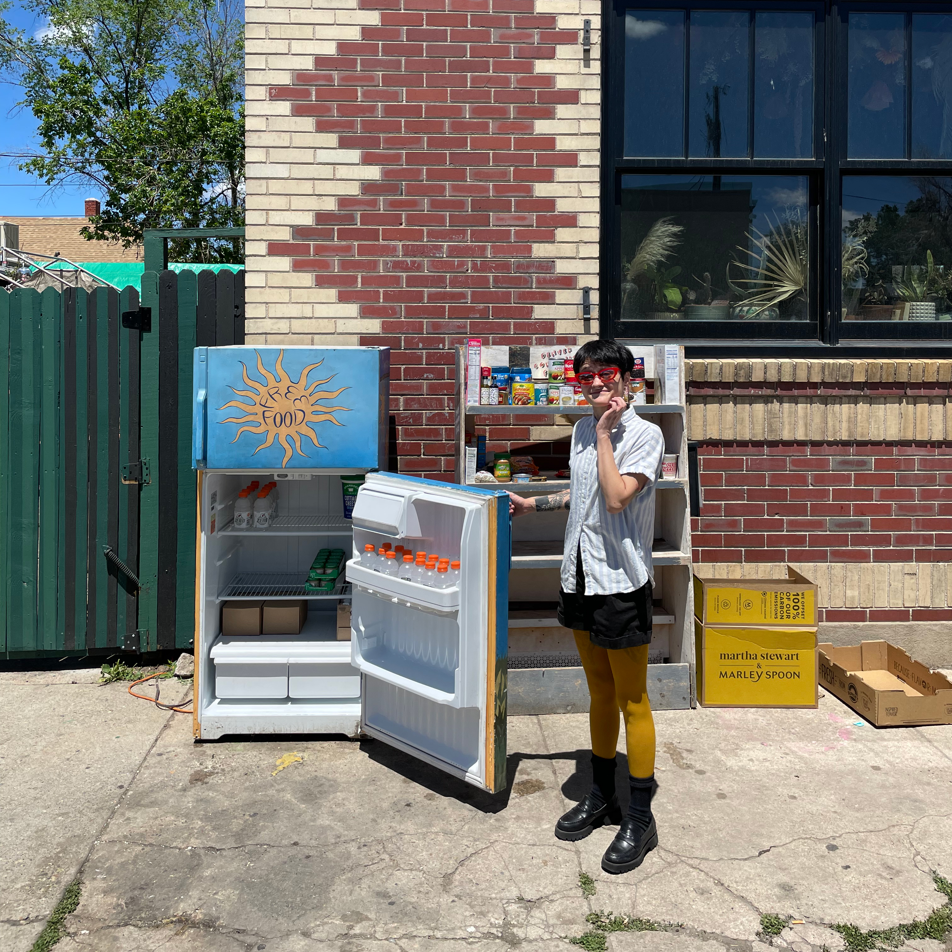 Person standing next to a fridge that is painted and sitting in front of a brick building.