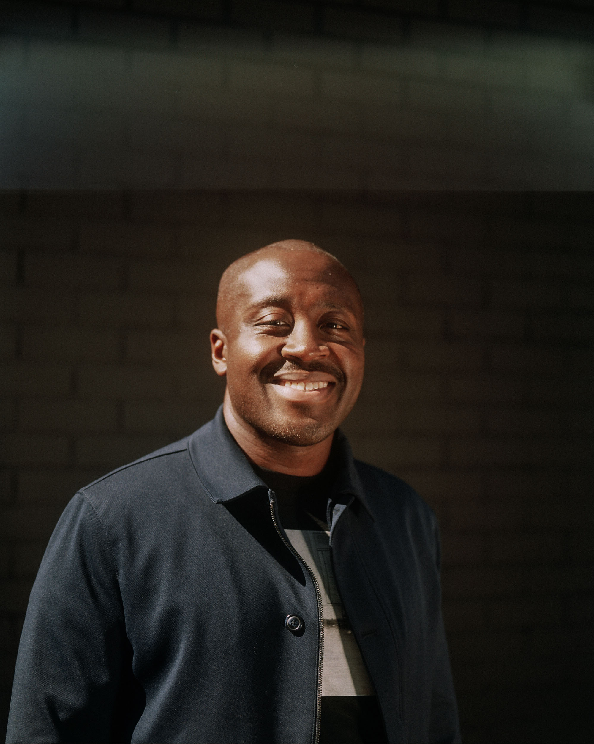 Portrait of curator Larry Ossei-Mensah standing in front of a black wall. He has a big smile on his face, and is sporting a casual collared coat and stubble on his face. 
