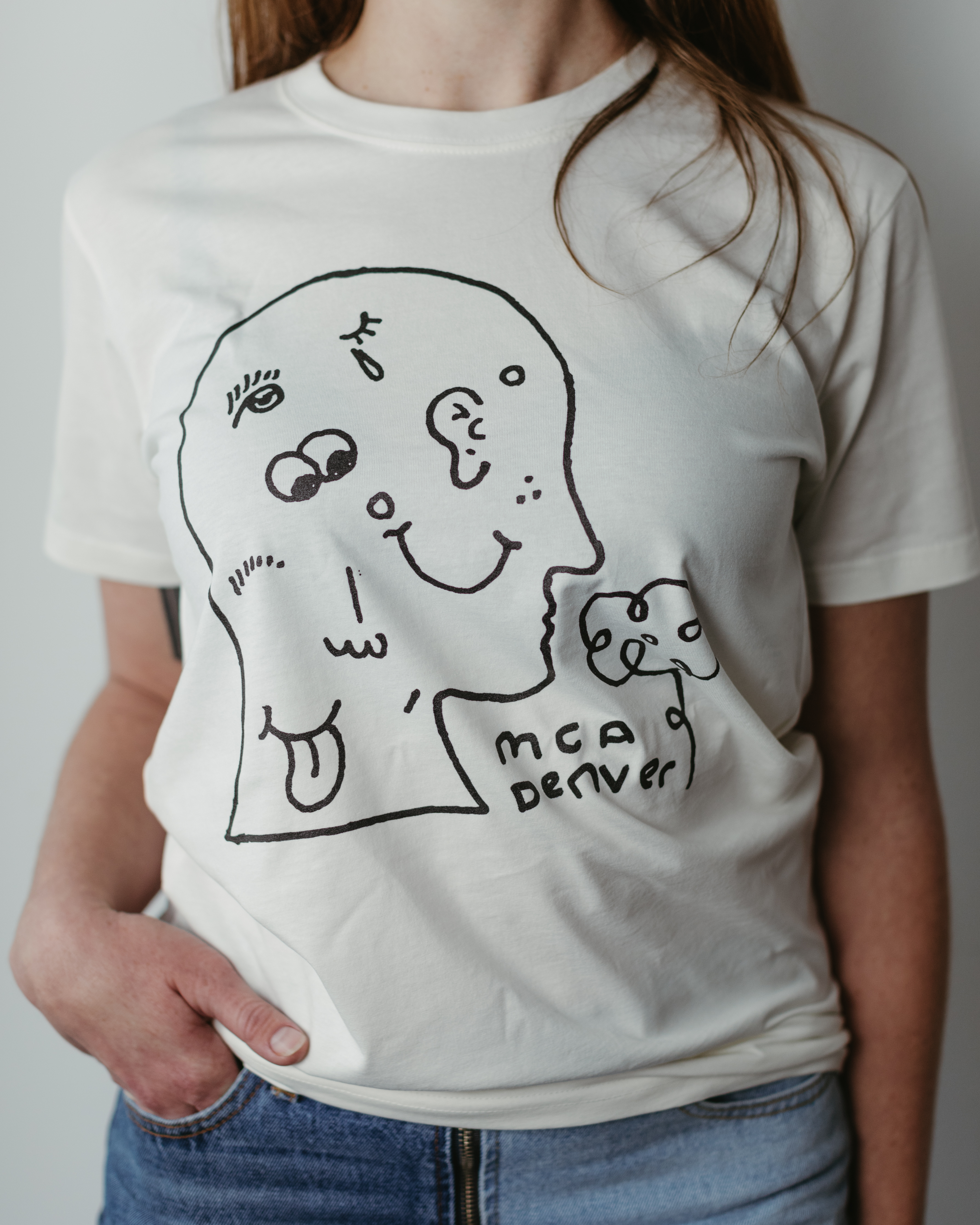 A close up image of a person wearing a white t-shirt with a black and white line drawing of the outline of a human profile on it. There is a small flower near the nose, which makes it look as though the figure on the shirt is smelling the flower. There is text between the flower and the head that reads “MCA Denver”. Inside the head are eyes, an ear, a smiley face, a mouth with a tongue sticking out, and a few other facial features.