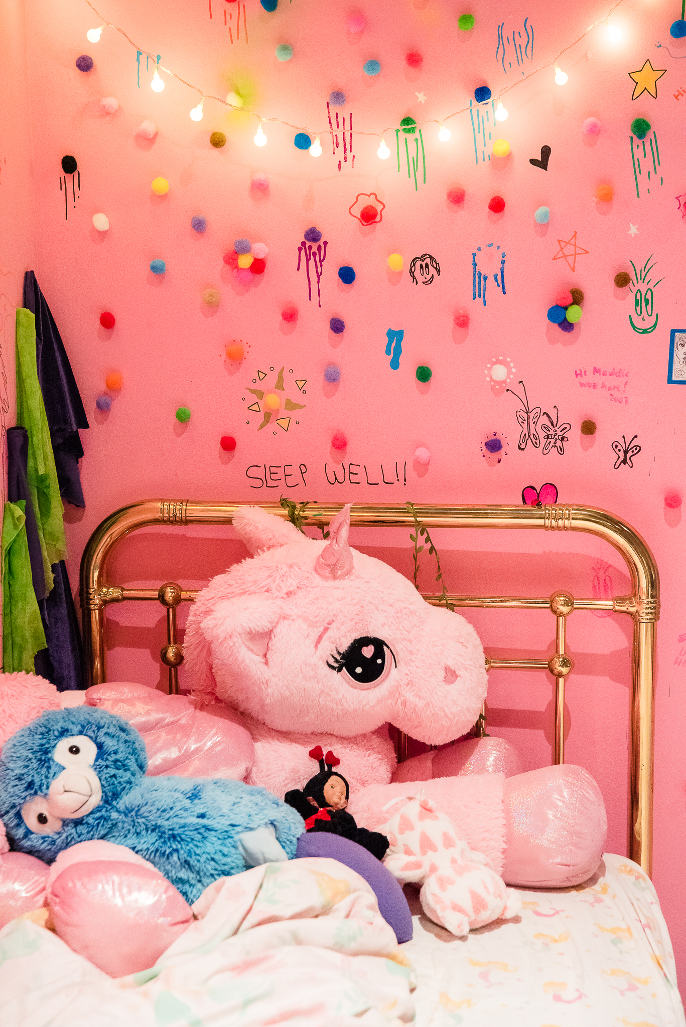  Corner of a gallery space that is bright pink. There are little colorful pom poms and string lights affixed to the wall. In view, there’s a pink unicorn laying in a bed with a gold bed frame. Above the bed frame on the wall is text that reads, “sleep well!!”