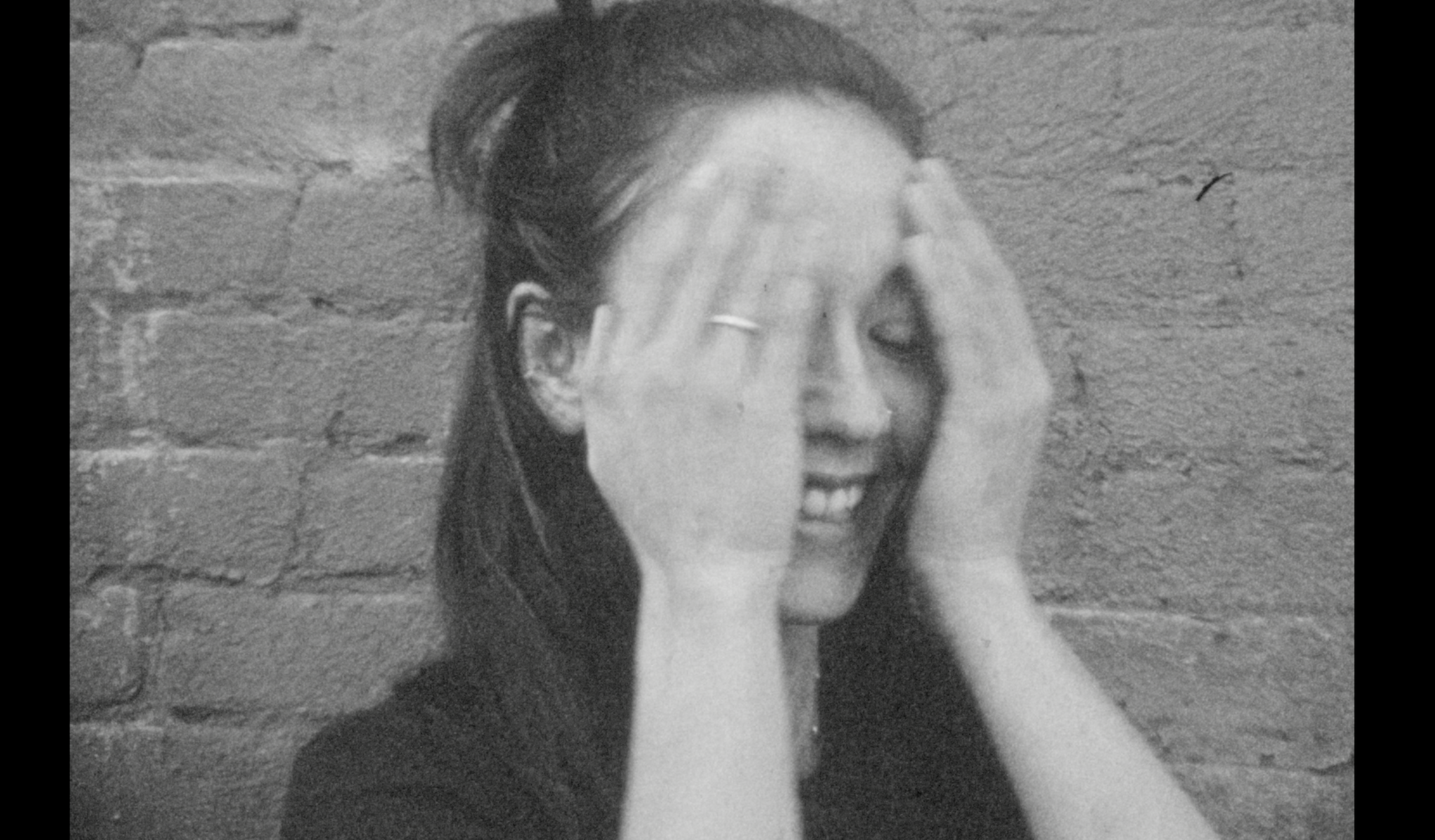 Portrait of Lucia in black and white. Lucia is looking to her left, smiling, and cover her face with her hands