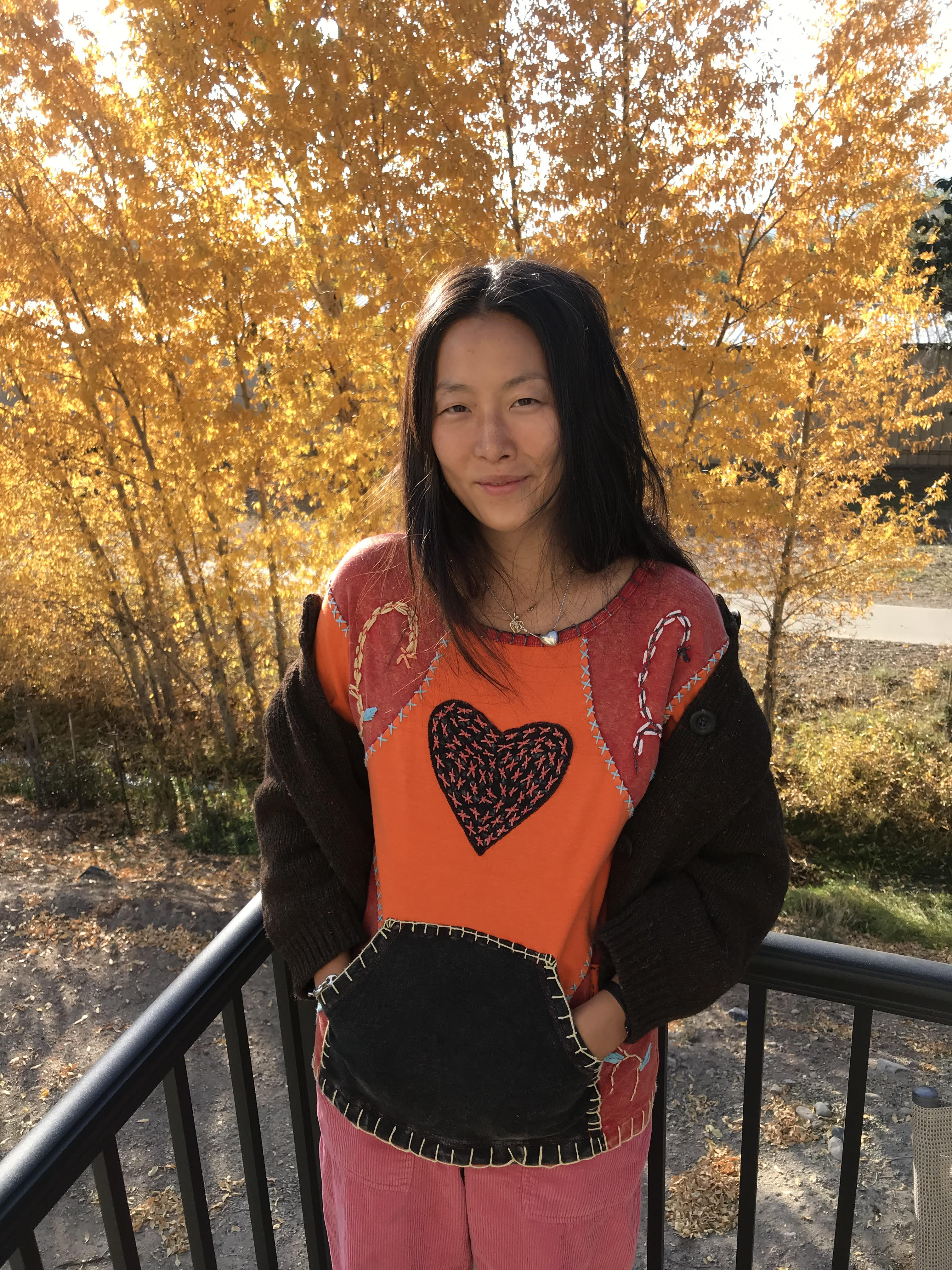 Portrait of artist Maia Ruth Lee wearing an orange and black hooded sweatshirt with bright orange leaves in the background. 