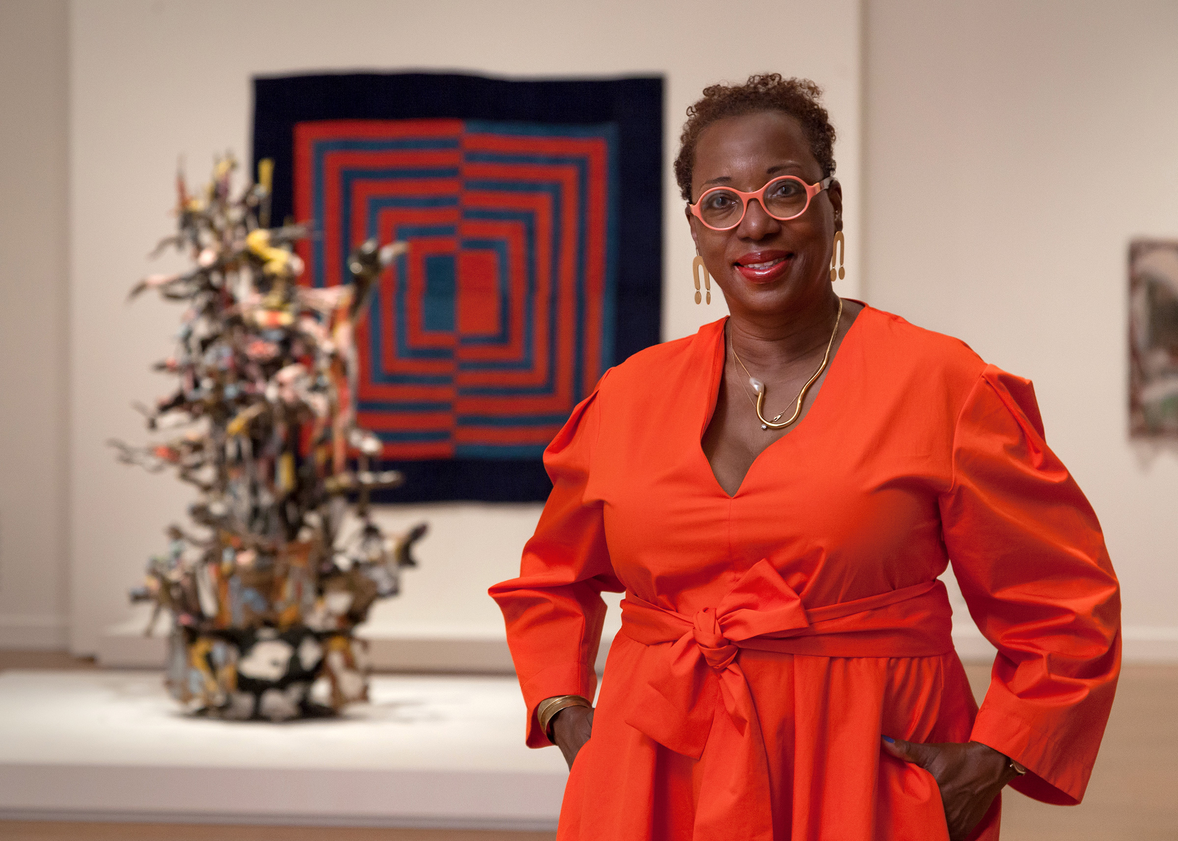 Curator Valerie Cassel Oliver sporting a red dress, gold necklace and earrings, and circular glasses with light red frames. She is standing in front of an artwork on the wall and sculpture on the ground.