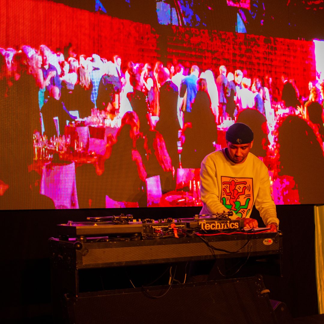 DJ performing in front of a screen displaying attendees at a Gala, blanketed in a pink hue created by the overhead lights. 