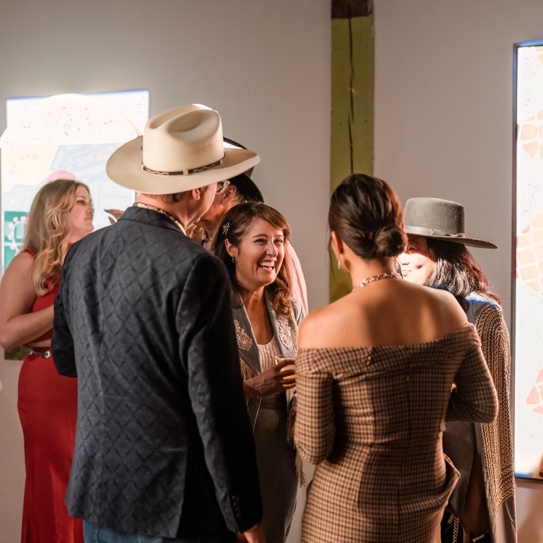 People standing in a gala talking and smiling. Artwork is hung up on walls behind them.