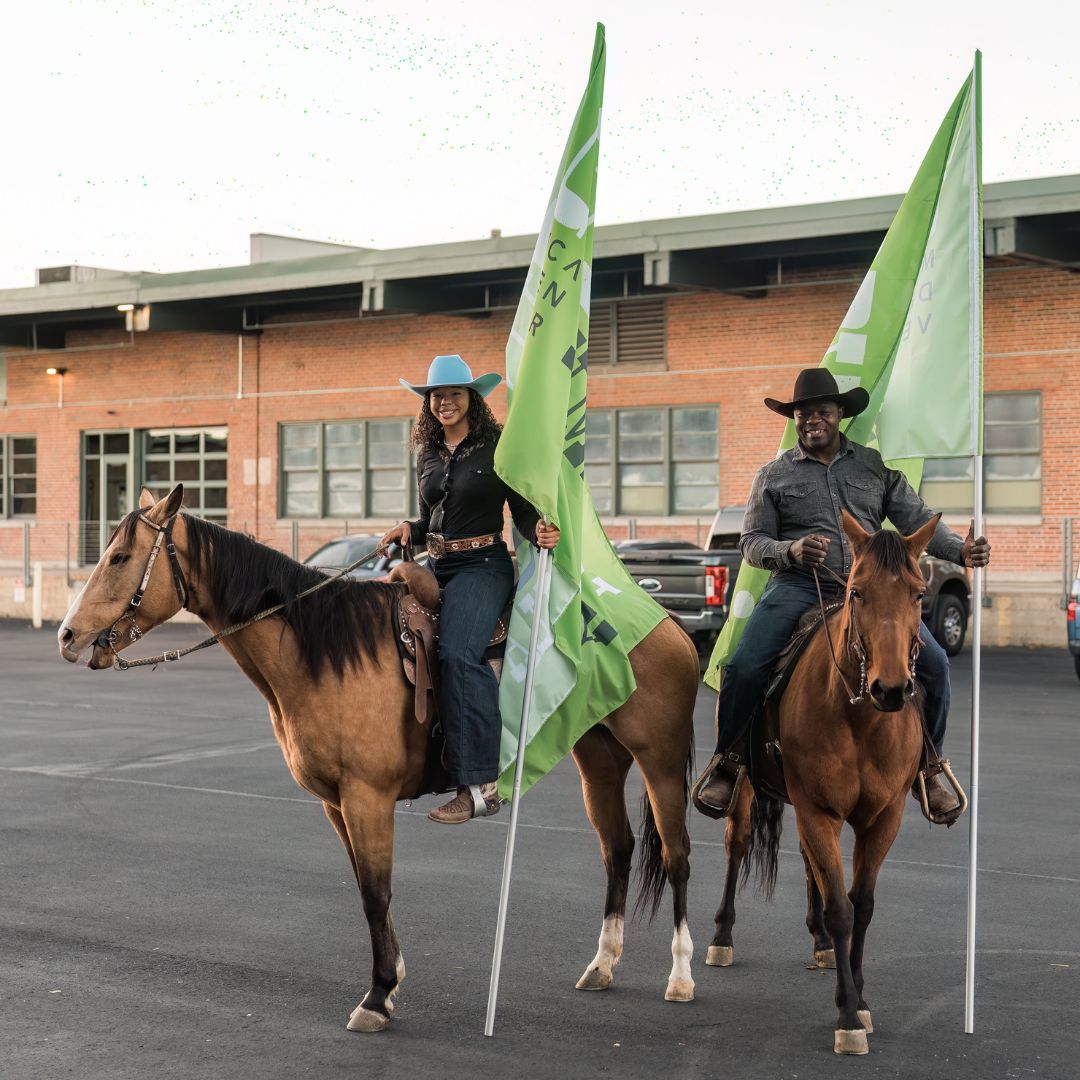 Two people riding horses, holding green flags.