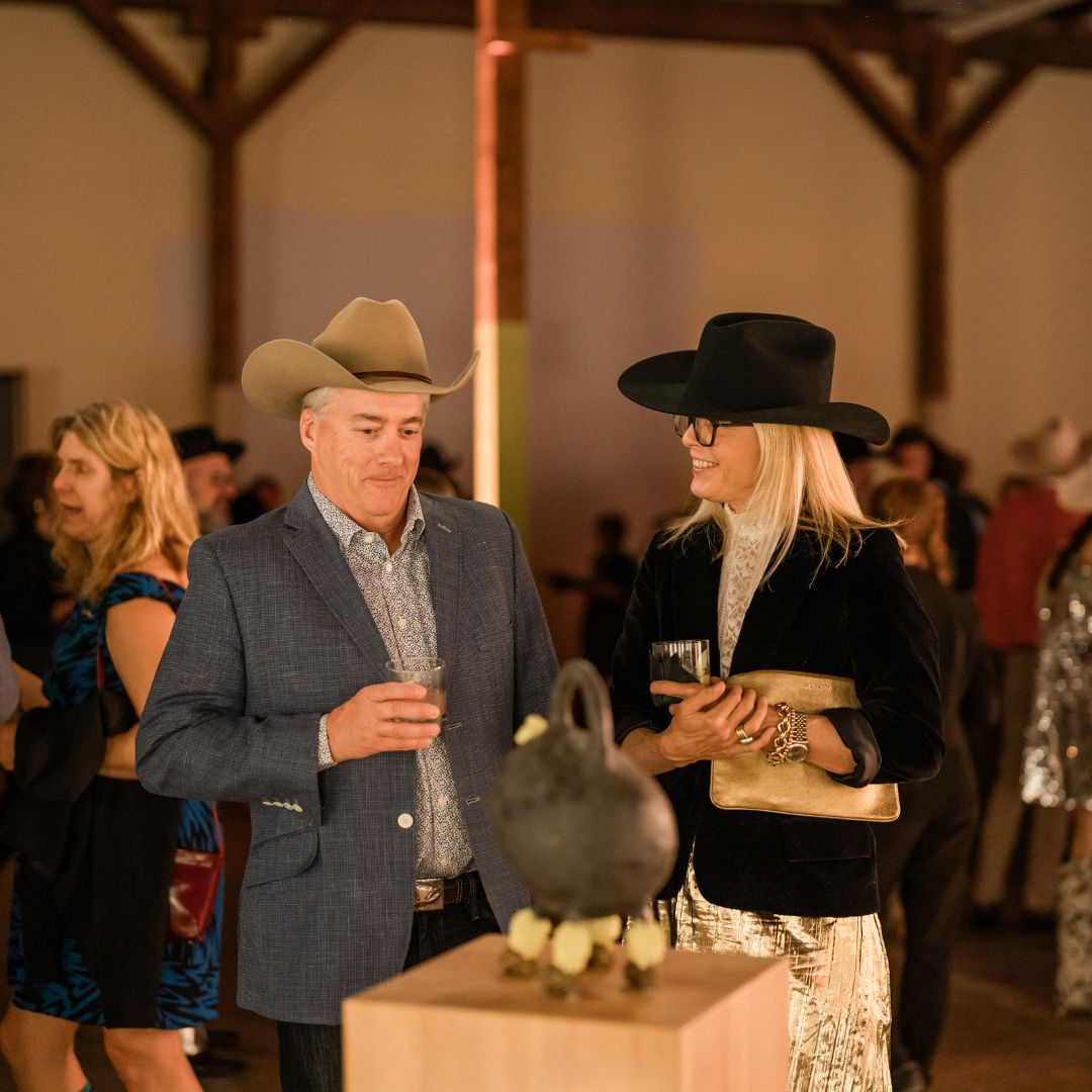 People standing in a gala, wearing cowboy hats and looking at a sculpture on a pedestal.