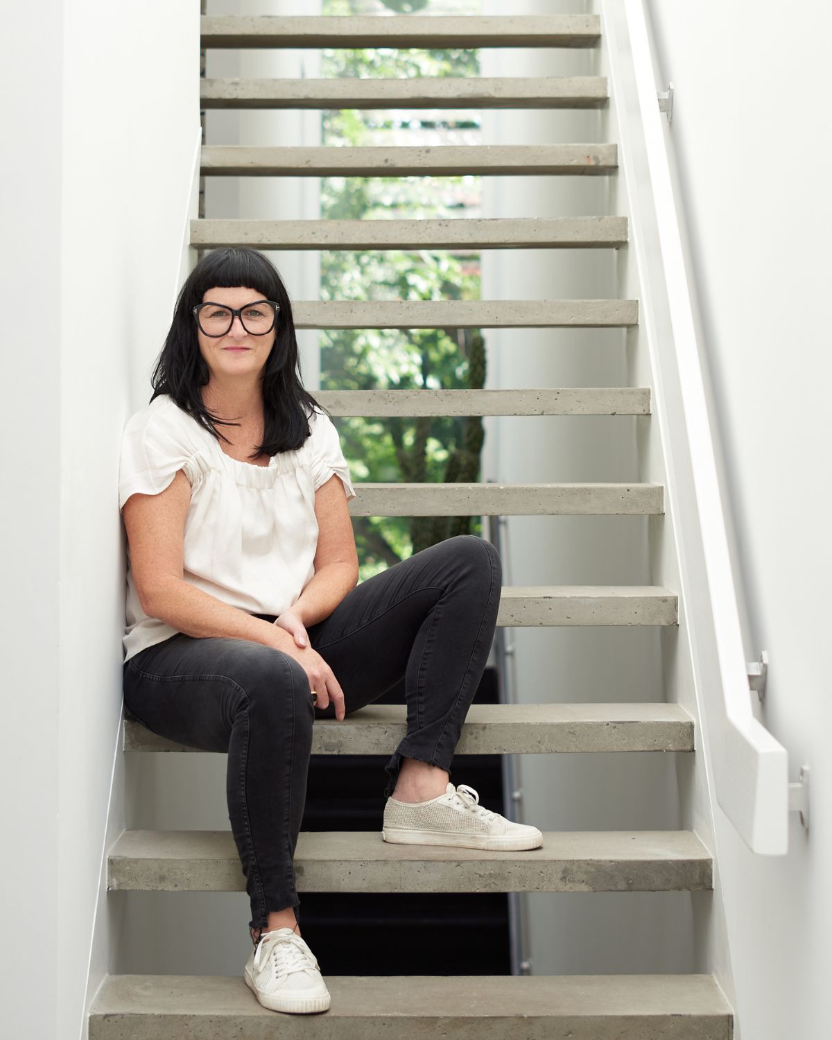 Artist Tara Donovan sporting black hair, big glasses, black pants, and a white shirt. She is seated on a flight of stairs.