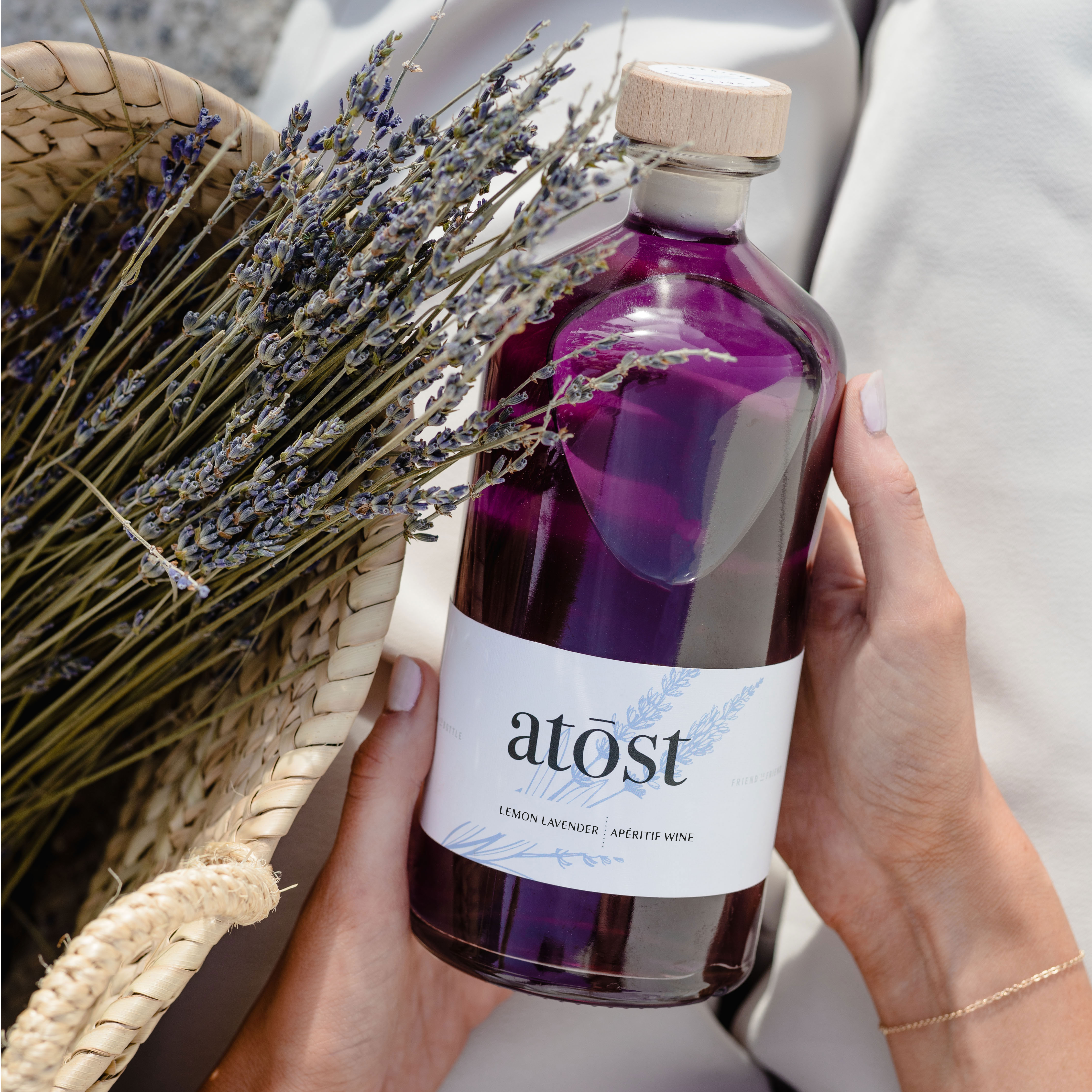 an image of the atost lemon lavender aperitif that is a beautiful deep purple color, held in hands and on a lap of white silk linen next to a weaved basket with dried lavender in it. 