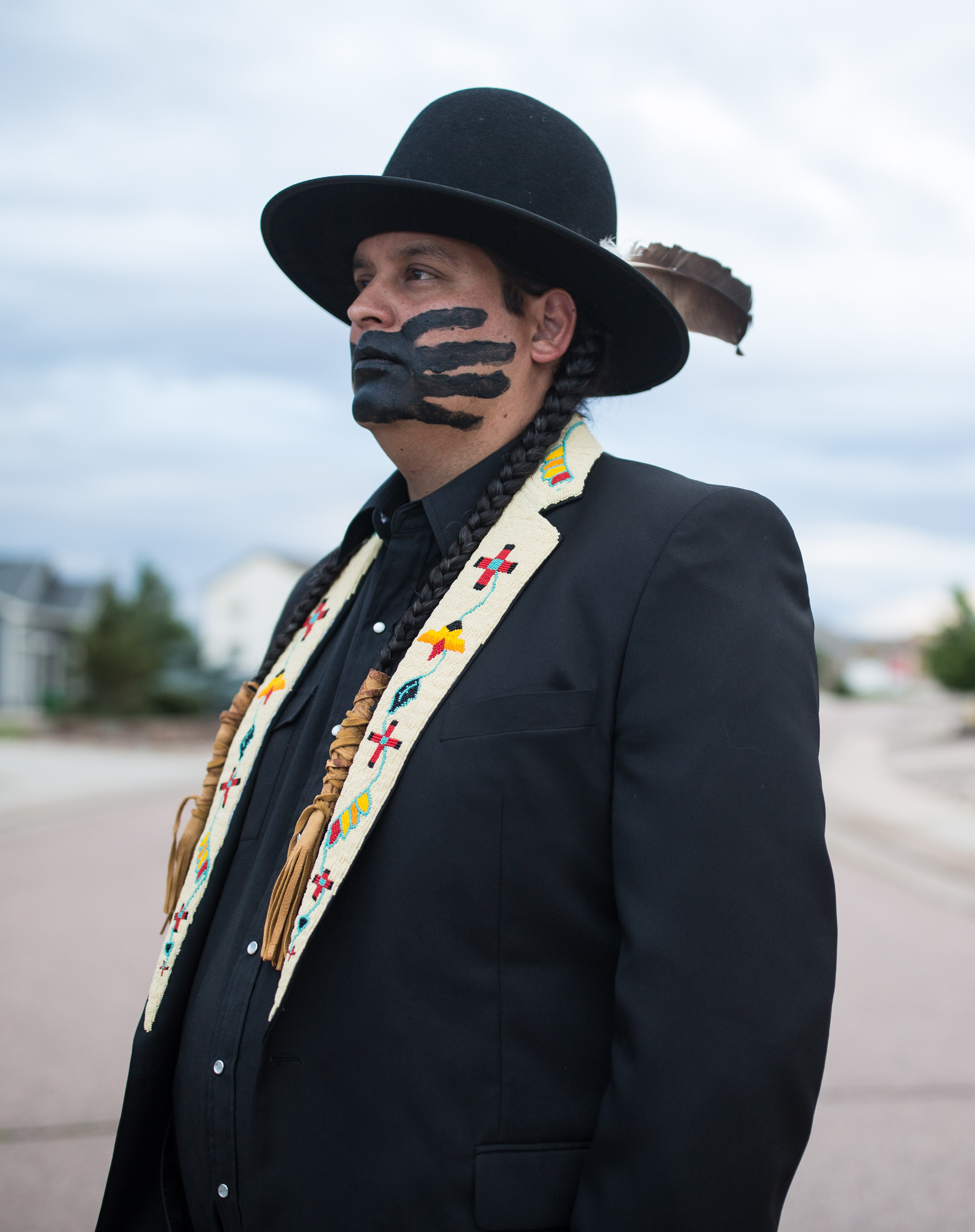 Image of artist Gregg Deal. Gregg is wearing a black full brimmed hat, and a black suit with Native American style beading on the lapels of the jacket. Gregg has a black paint handprint on his face, covering his mouth.