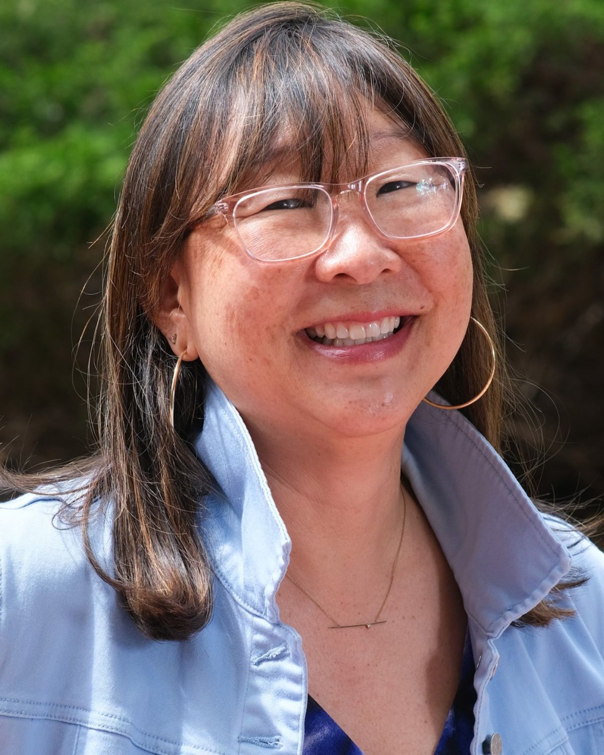 Headshot of Stacey Shigaya, standing outside and sporting bangs, clear glasses, and a popped collar. 