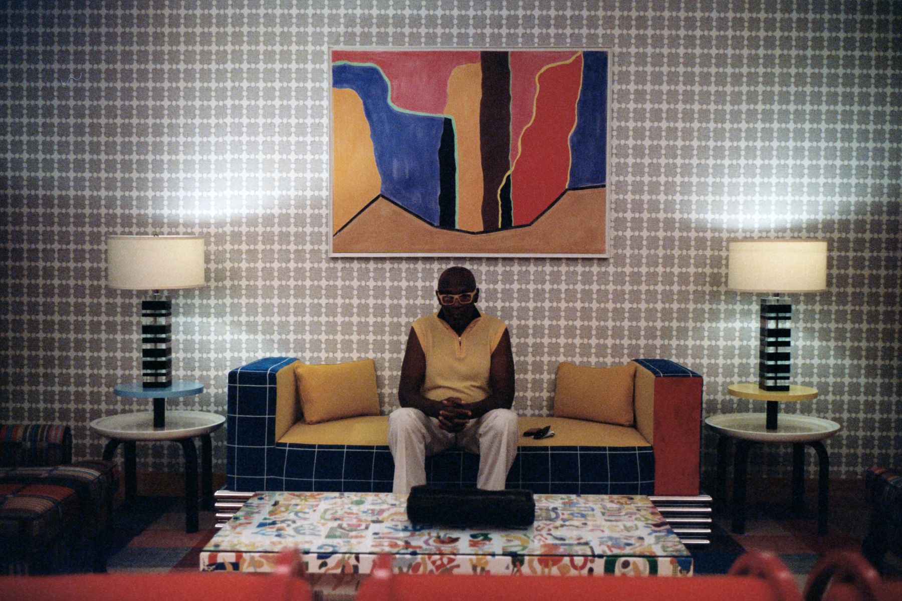 Person sitting on a couch in a retro room, with their hands to themselves. There is a big abstract artwork directly above them on the couch.