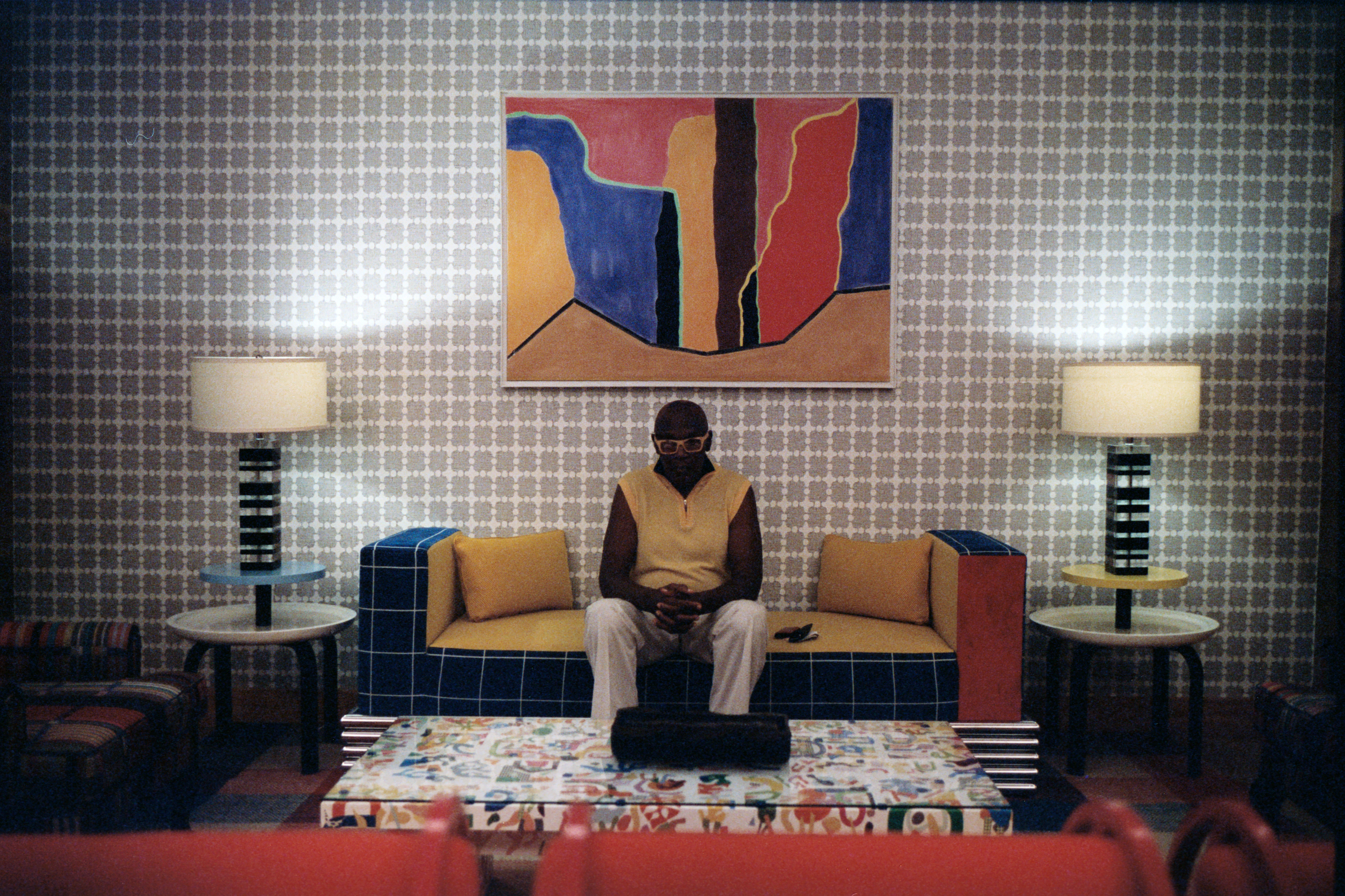 Person sitting on a couch in a retro room, with their hands to themselves. There is a big abstract artwork directly above them on the couch.