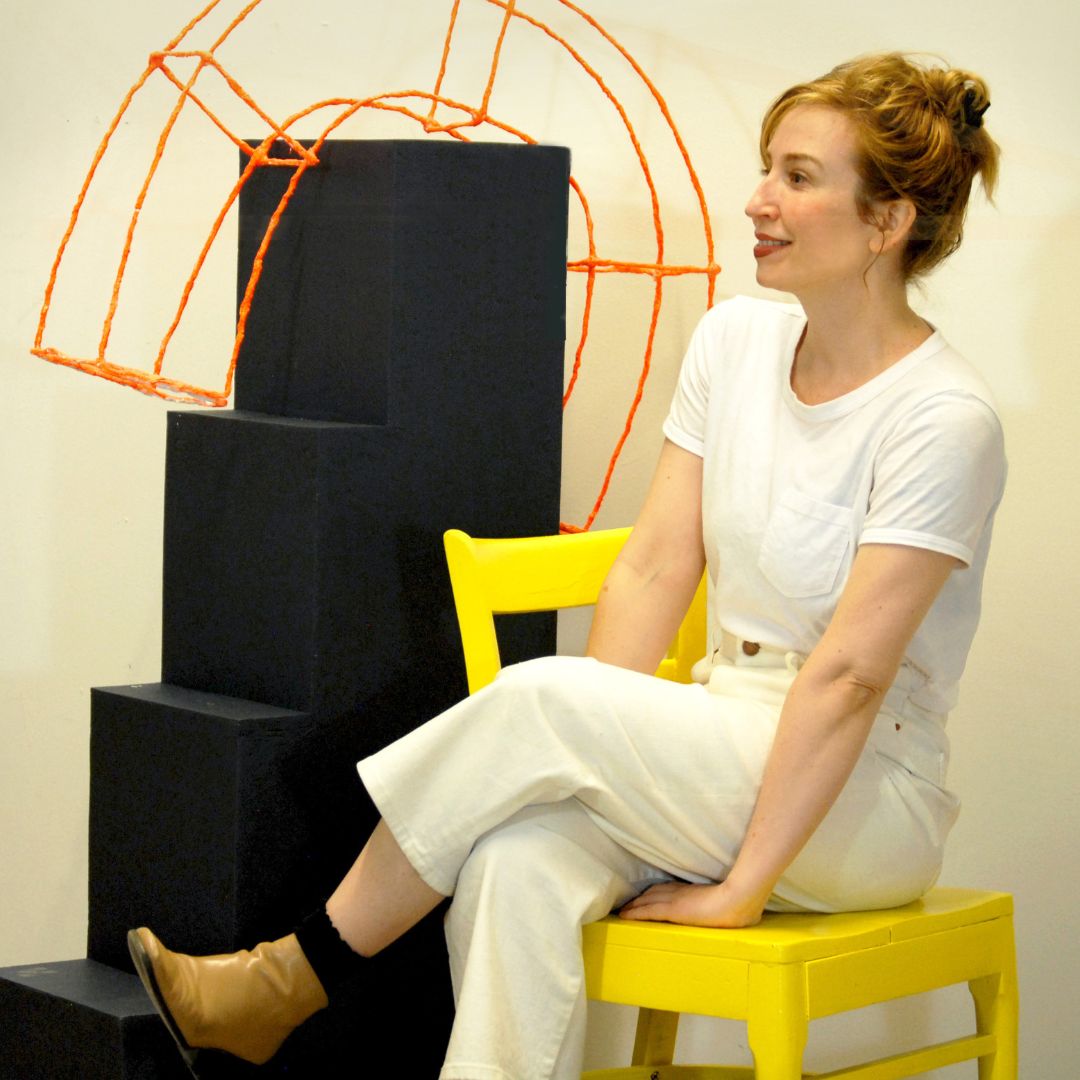 Portrait of artist Alicia Ordal, wearing all white, sitting on a yellow chair, and sitting next to what looks like a small black staircase.
