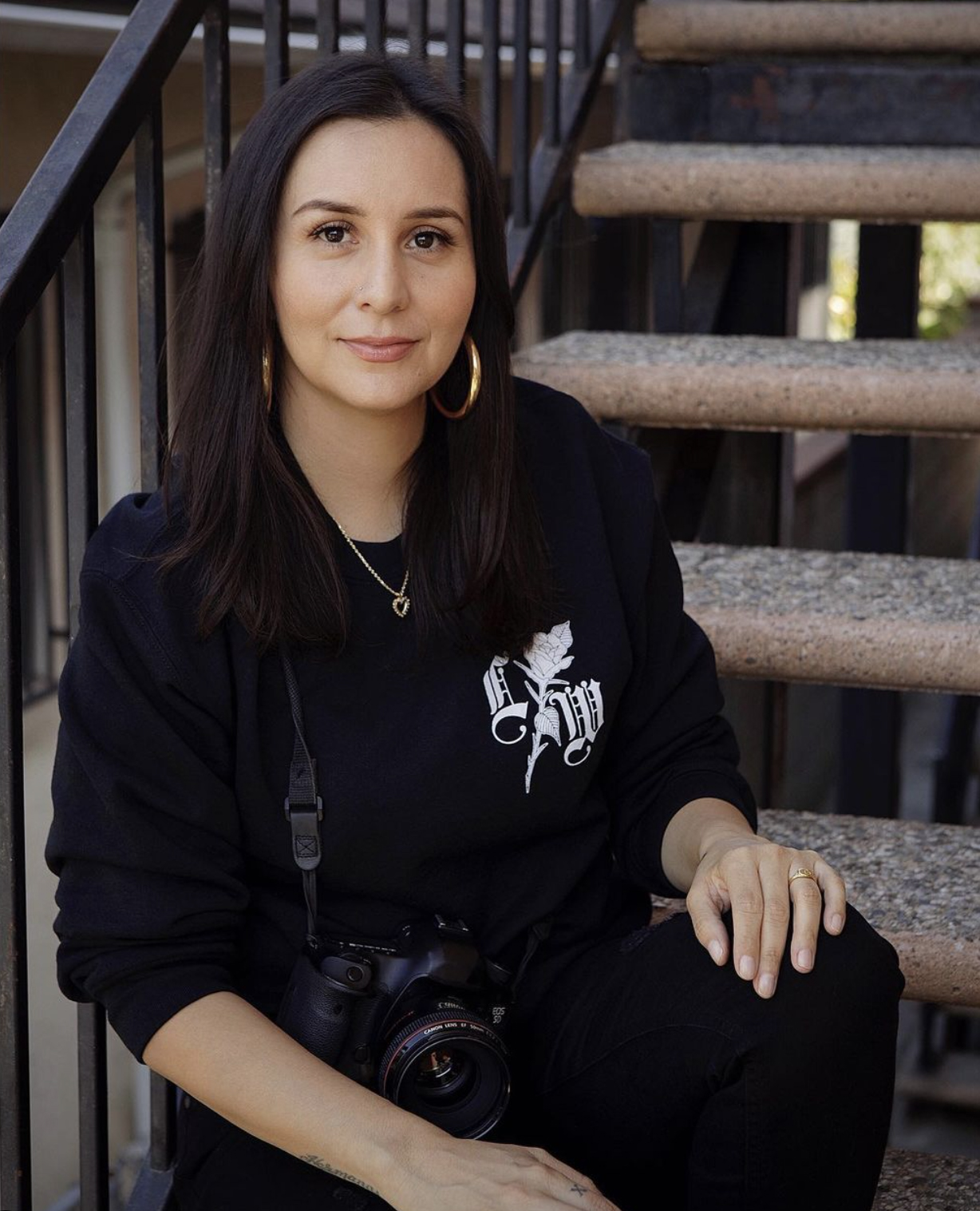 Image of photographer Amanda Lopez sitting on a staircase wearing all black with her camera hanging from her neck. 