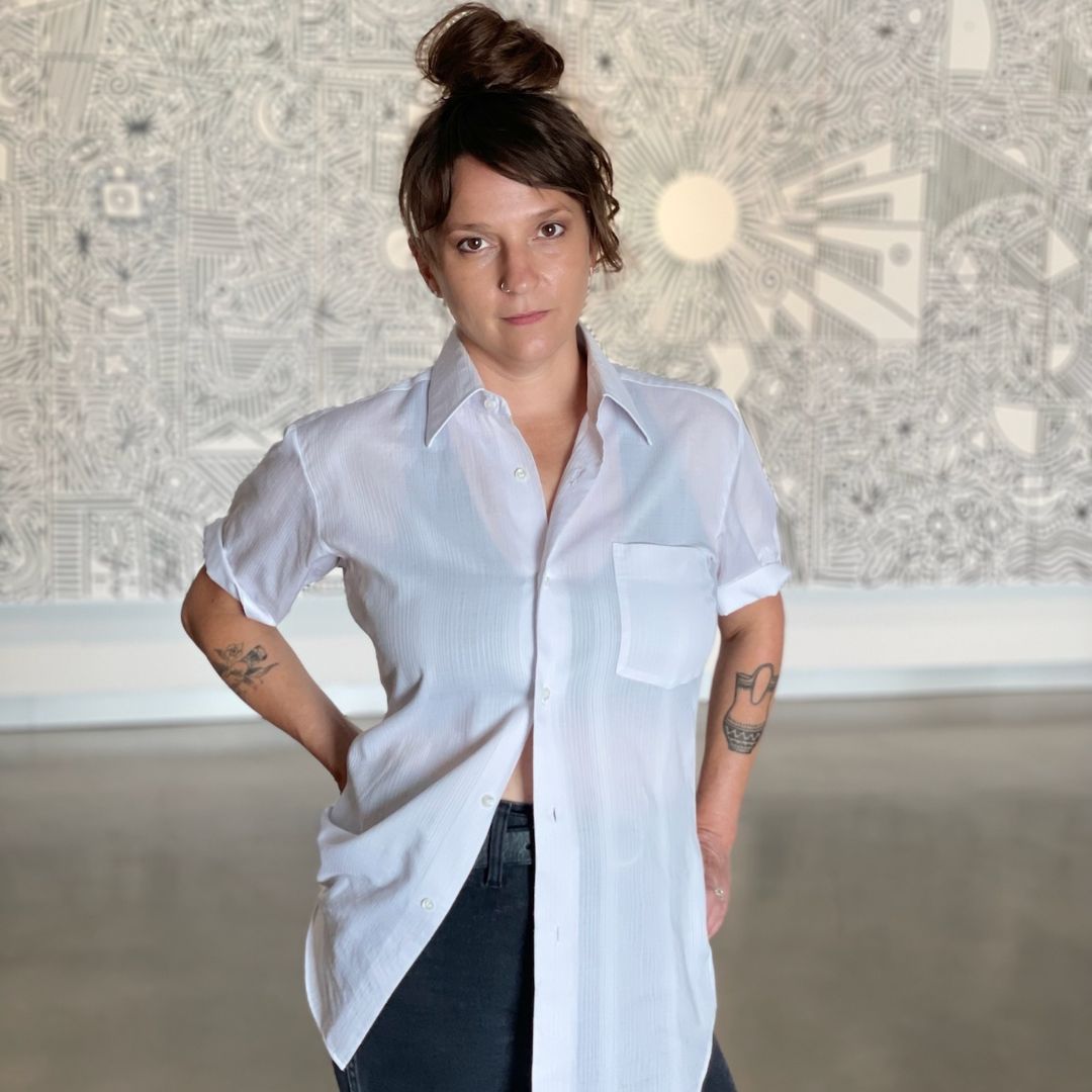 Portrait of artist Amber Cobb, sporting an up-do and a white short sleeve button down that reveals tattoos on her arms. 