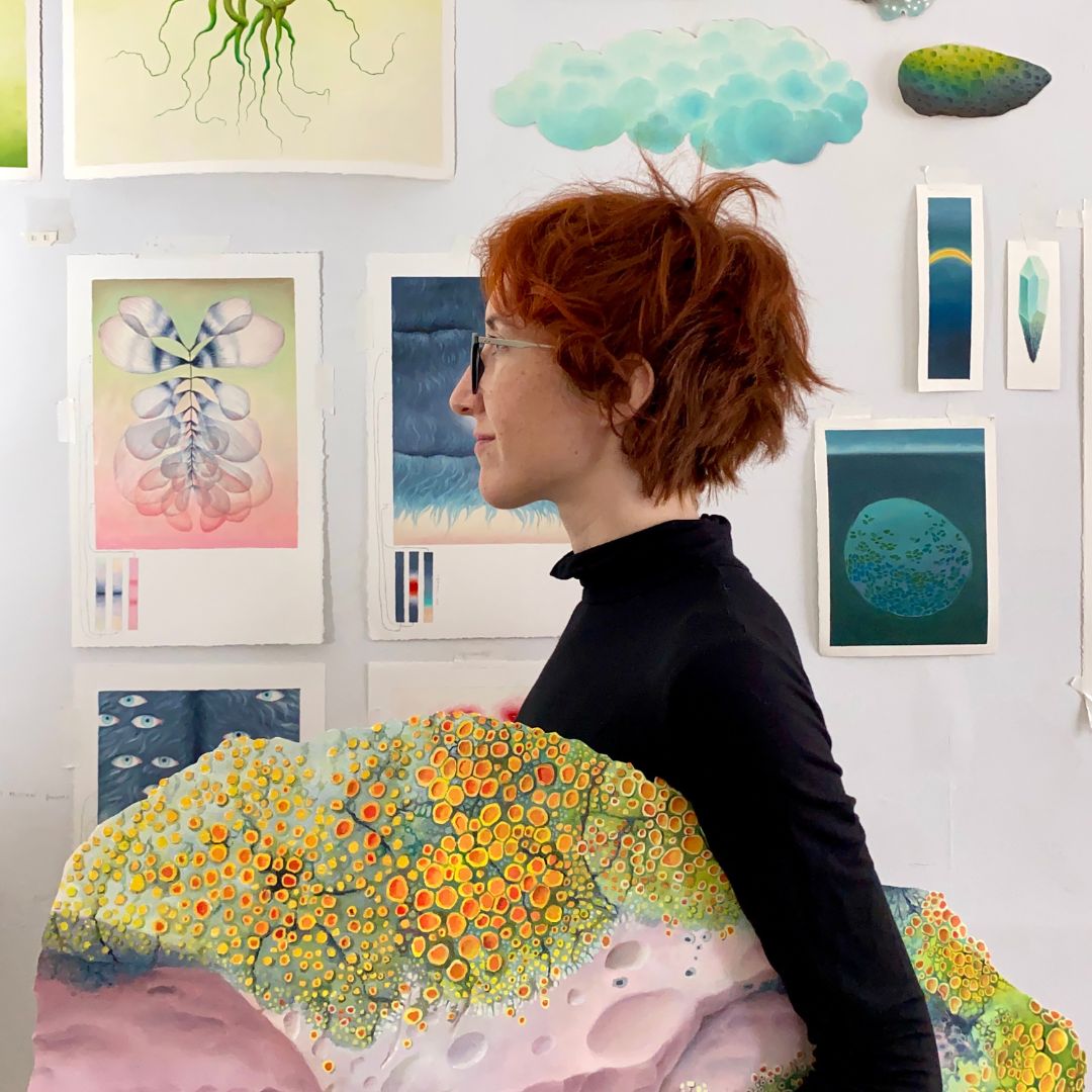 Portrait of Ashley Eliza Williams’s profile. Ashley is holding what looks like a large, colorful rock, and is standing in front of colorful artwork hung on the wall. 