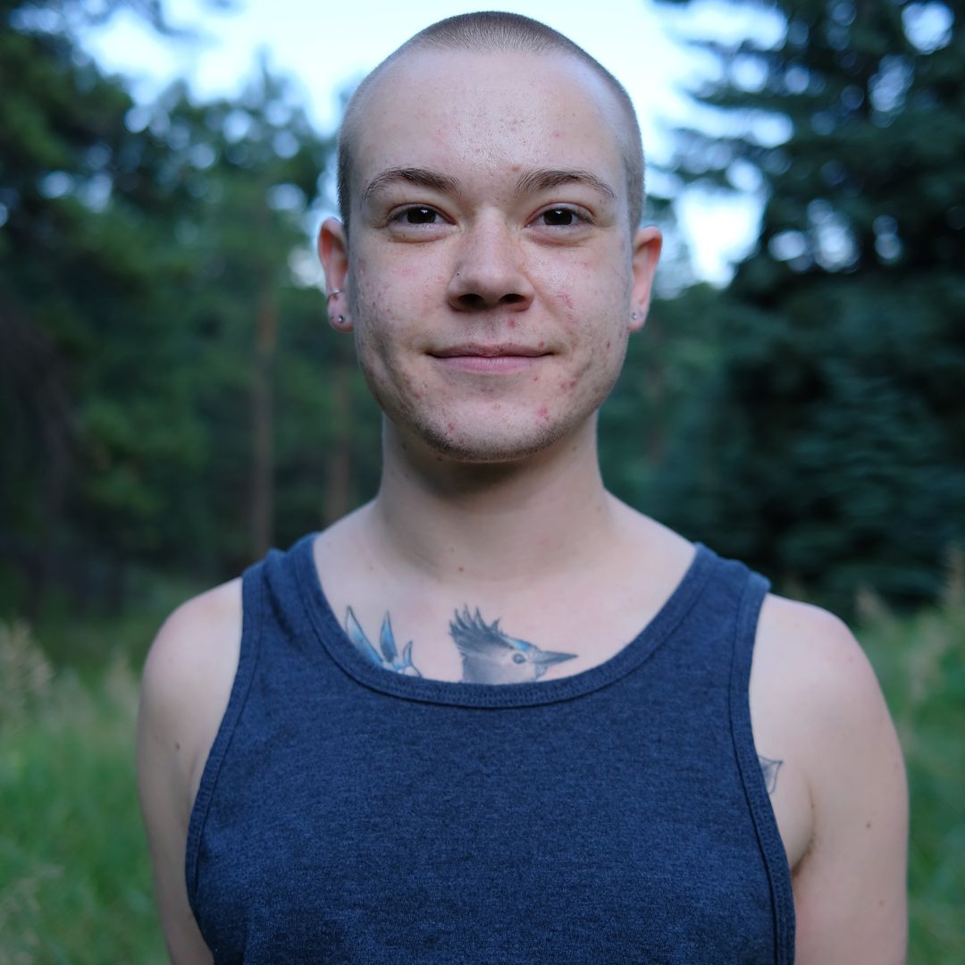 Person named Blue Lane sporting a gray tank top. They are standing in front of trees and grass, and smiling softly.