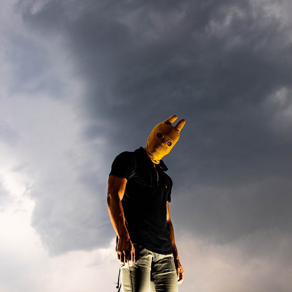 Band photo featuring one person standing with a mask over their face, posing in front of a stormy sky.