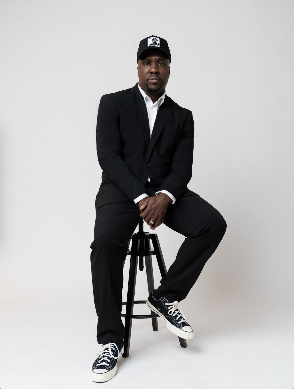 Image of musician Derrick Hodge wearing a black suit and black baseball cap. Derrick is sitting on a black stool in a white studio space. 