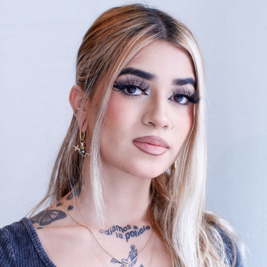 Portrait of a person named Destany Rodriguez, sporting dark eye makeup, a v neck shirt that reveals her tattoos, and a hair style where half her hair is pulled back.