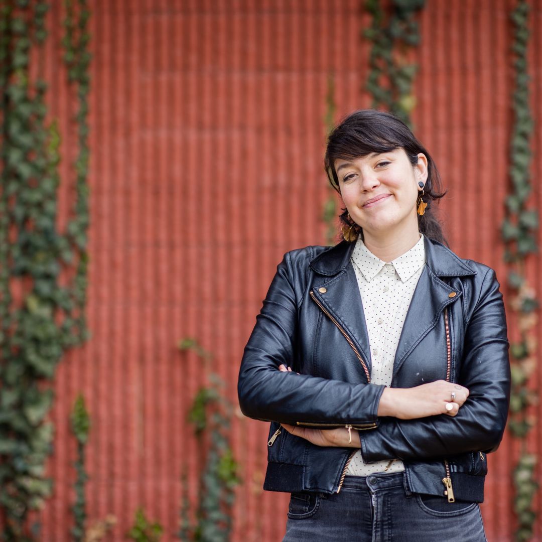 Portrait of artist Eileen Roscina, posing in front of a red wall covered in vines and sporting a black leather jacket and polka dot button down underneath. 