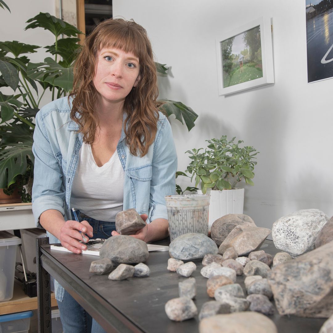 Portrait of artist Gretchen Marie Schaefer, leaning over a table covered in rocks. 