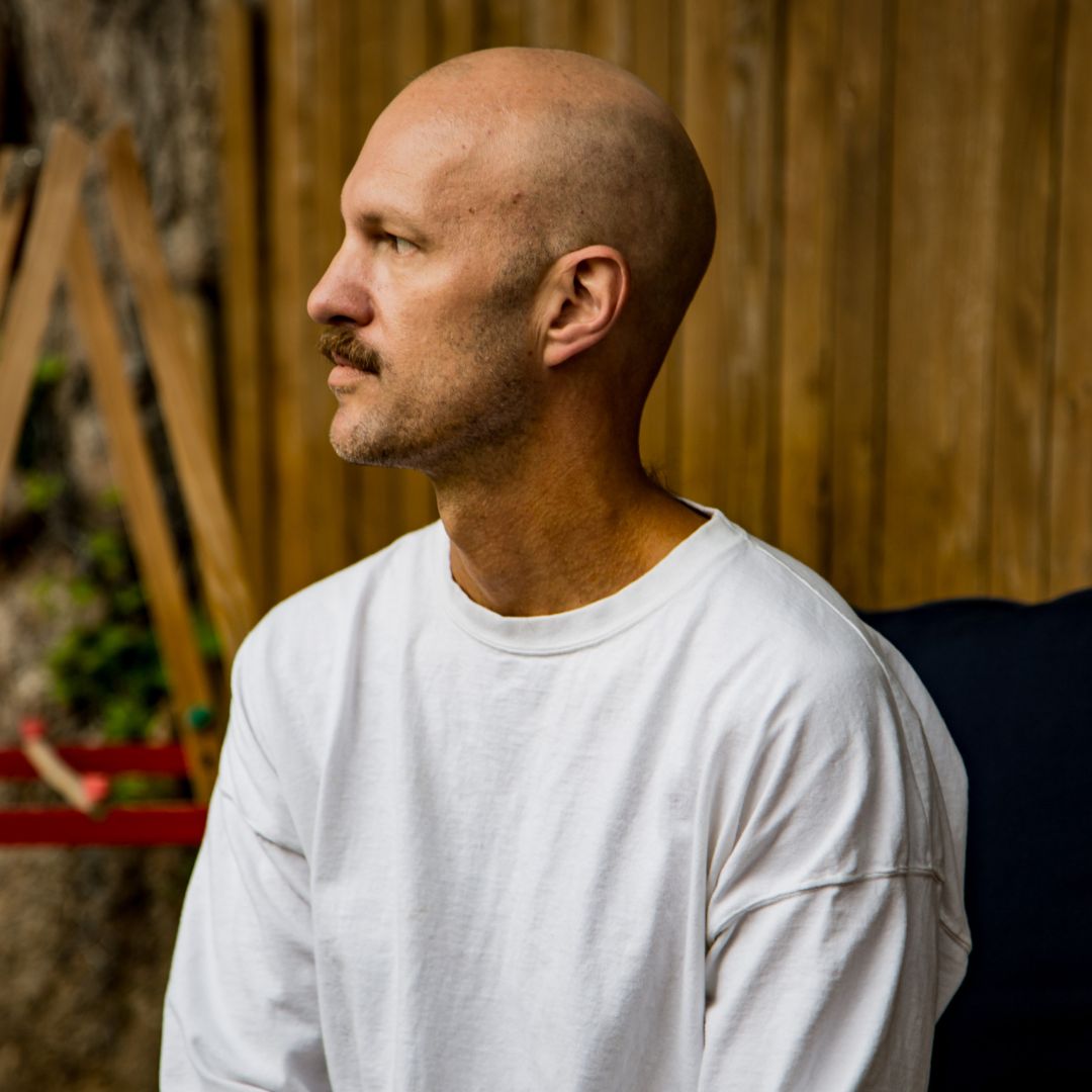 Portrait of artist Jeff Page, sporting a white t-shirt and mustache. Jeff is looking in another direction. 