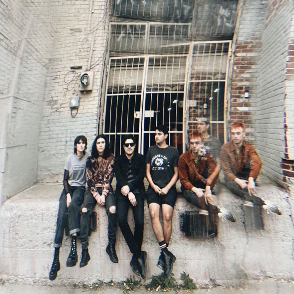 Band photo featuring six people sitting in what looks like an alleyway in front of brick walls.