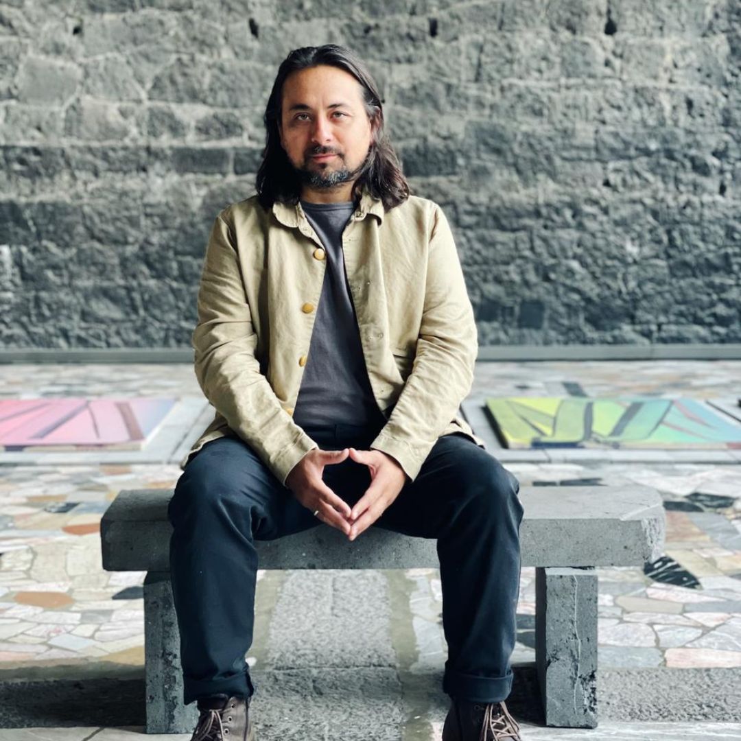 Portrait of artist Mario Zoots sitting on a bench, in front of a stone wall. Mario is sporting a black shirt, black pants, and a beige jacket. 