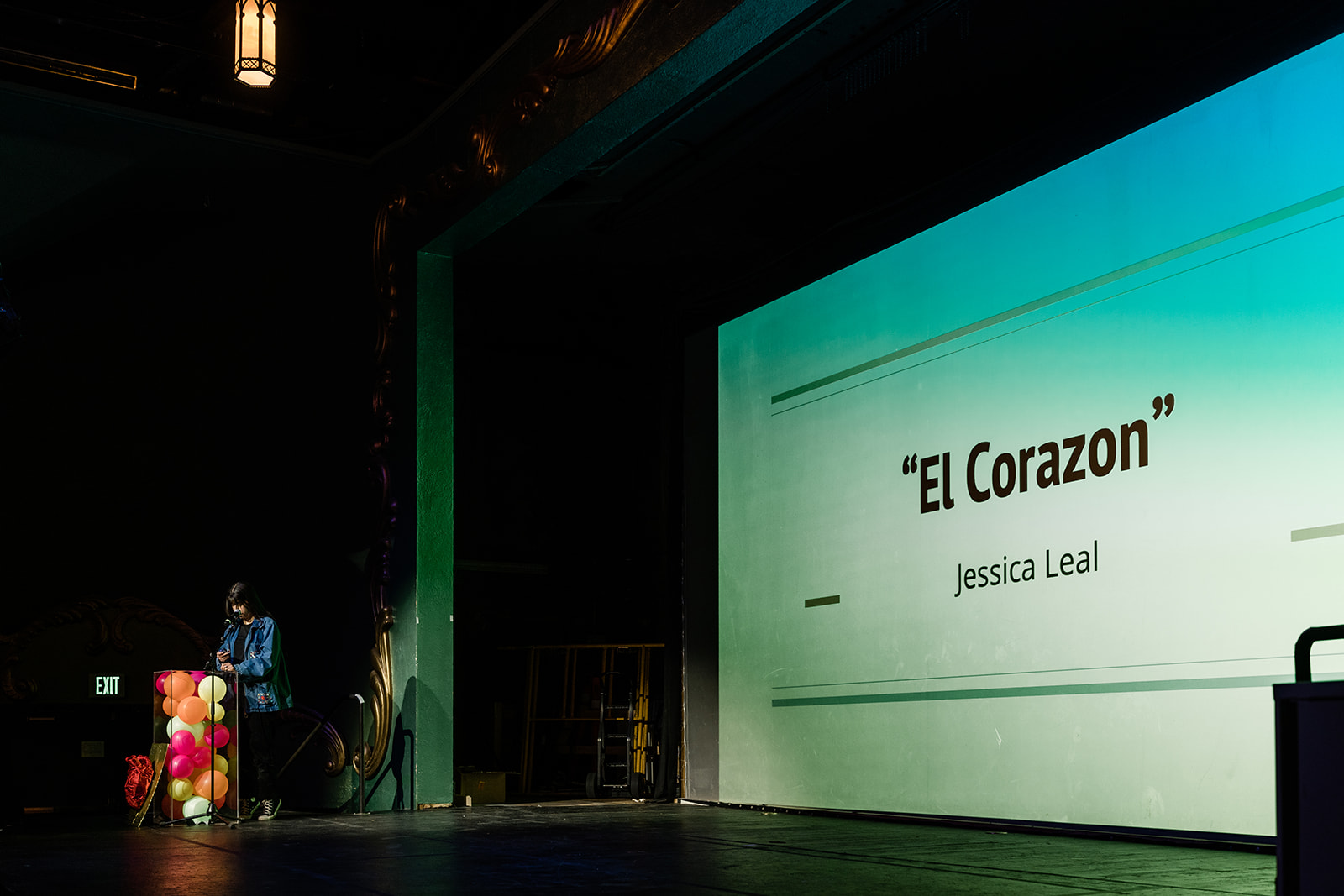 Photo of a young woman standing at a podium that is filled with balloons. She is onstage in front of a giant white screen that says "El Corazon" Jessica Leal