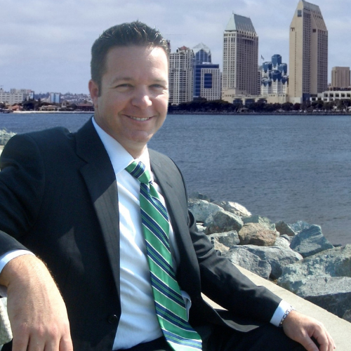 Portrait of Michael Bevis wearing a suit and striped tie, sitting next to a body of water and tall, city buildings behind him.