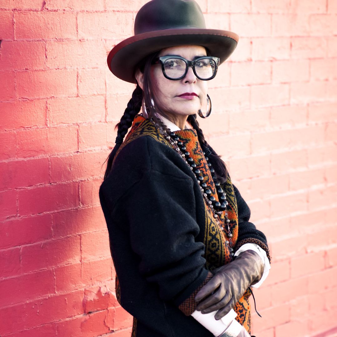 Designer named Mona Lucero standing in front of a red brick wall. Mona is sporting a hat, big glasses, and a layered outfit.