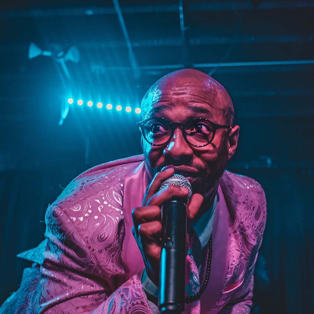 Band photo featuring a person singing in a microphone under neon lights.
