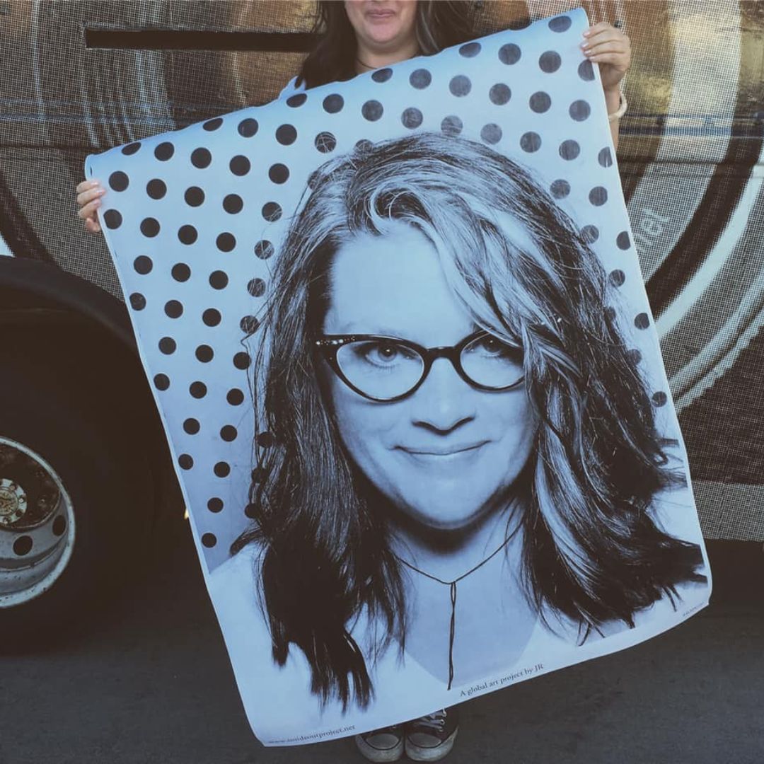Portrait of artist Rebecca Vaughan holding up a large portrait of herself. In the portrait she is sporting cat eye glasses and is smirking.