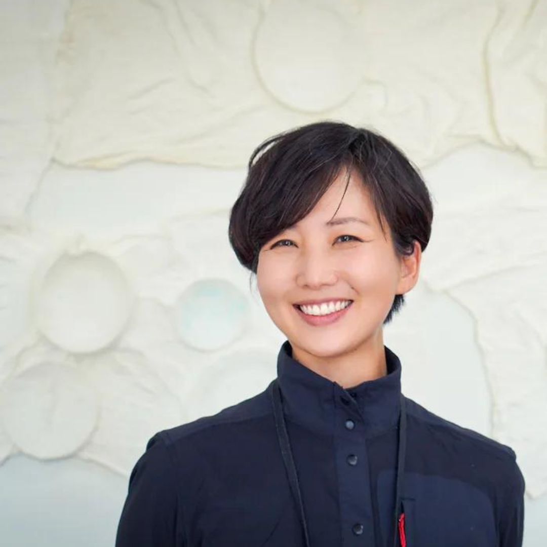 Portrait of artist Sammy Seung-Min Lee, sporting a big smile, jacket with a high neck, and standing in front of a white textured background. 