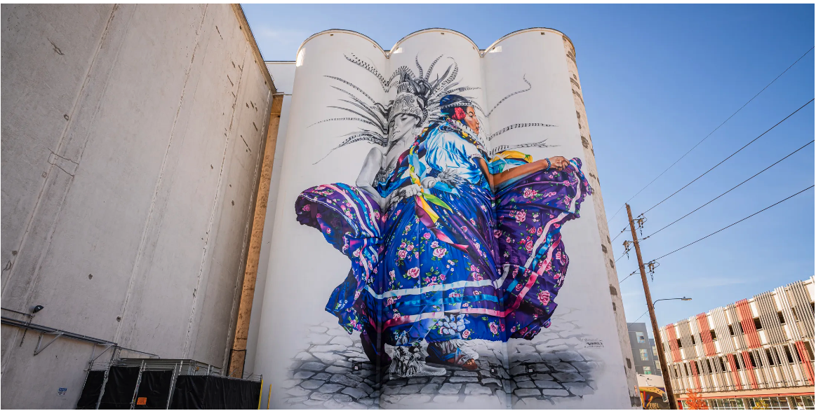 Bimmer Torres mural in Rino of a colorful dancer in aztecan traditional dress painted in the most vibrant of blues, purples, yellow and red, on the side of two huge grain silos. 